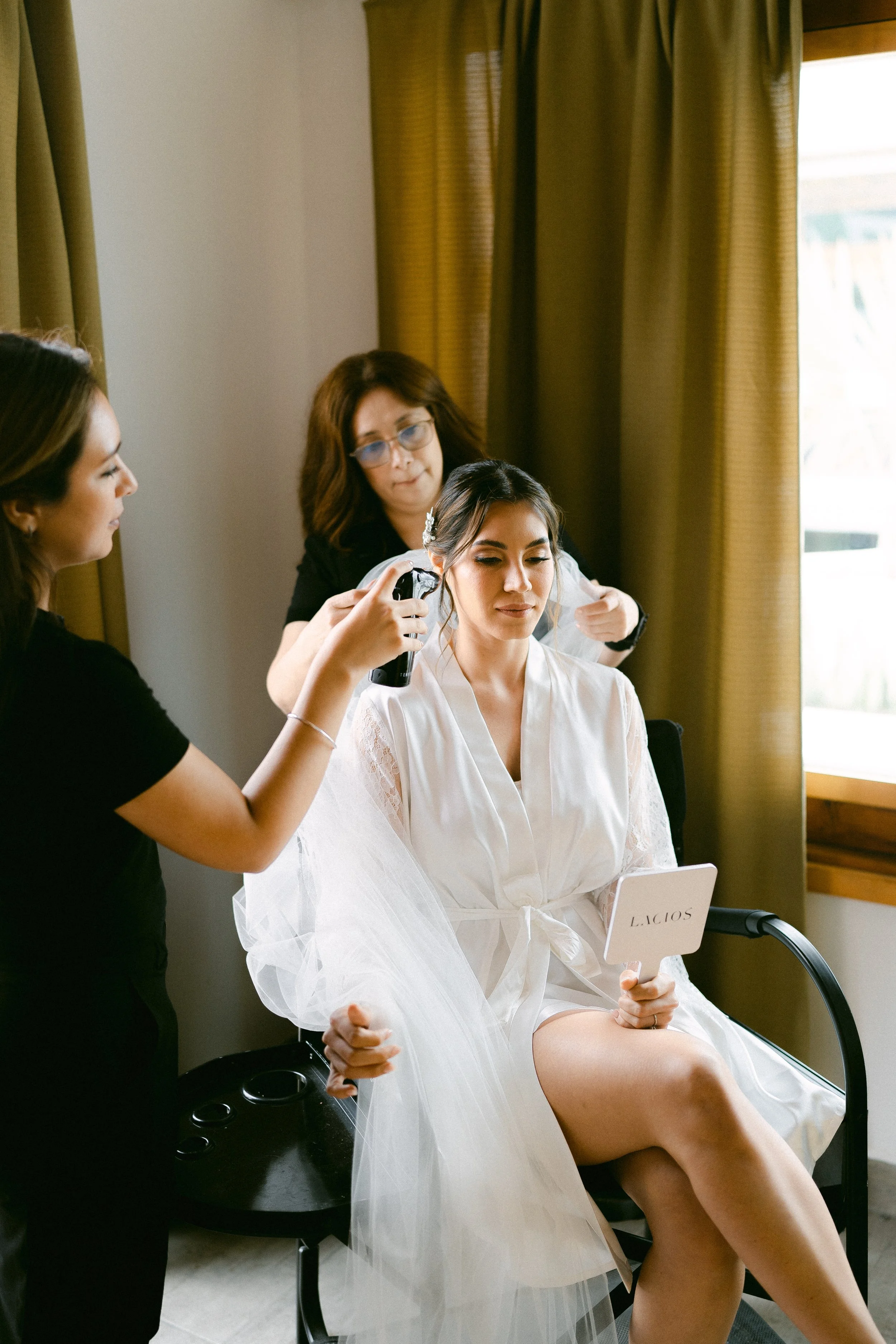 Una novia con bata blanca se sienta en una silla mientras una maquilladora en ensenada le aplica maquillaje en aerografo y otra mujer la peina el cabello en una habitación con cortinas verdes y una ventana con luz natural en valle de guadalupe