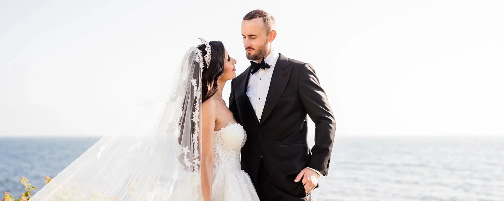 Pareja de novios en boda en la playa, la novia viste vestido blanco y velo, el novio traje negro y corbata de moño, abrazados frente al mar
