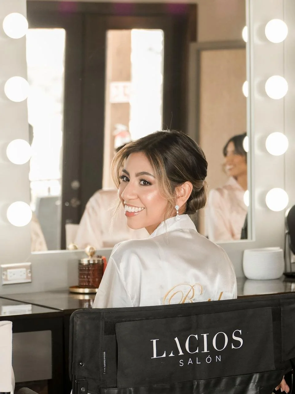 Una mujer sonriente en un salón de belleza frente a un espejo con luces. Ella lleva pendientes y una bata blanca con letras doradas en la parte trasera, y en la silla hay una bolsa negra con el logo 'LACIOS SALÓN'.