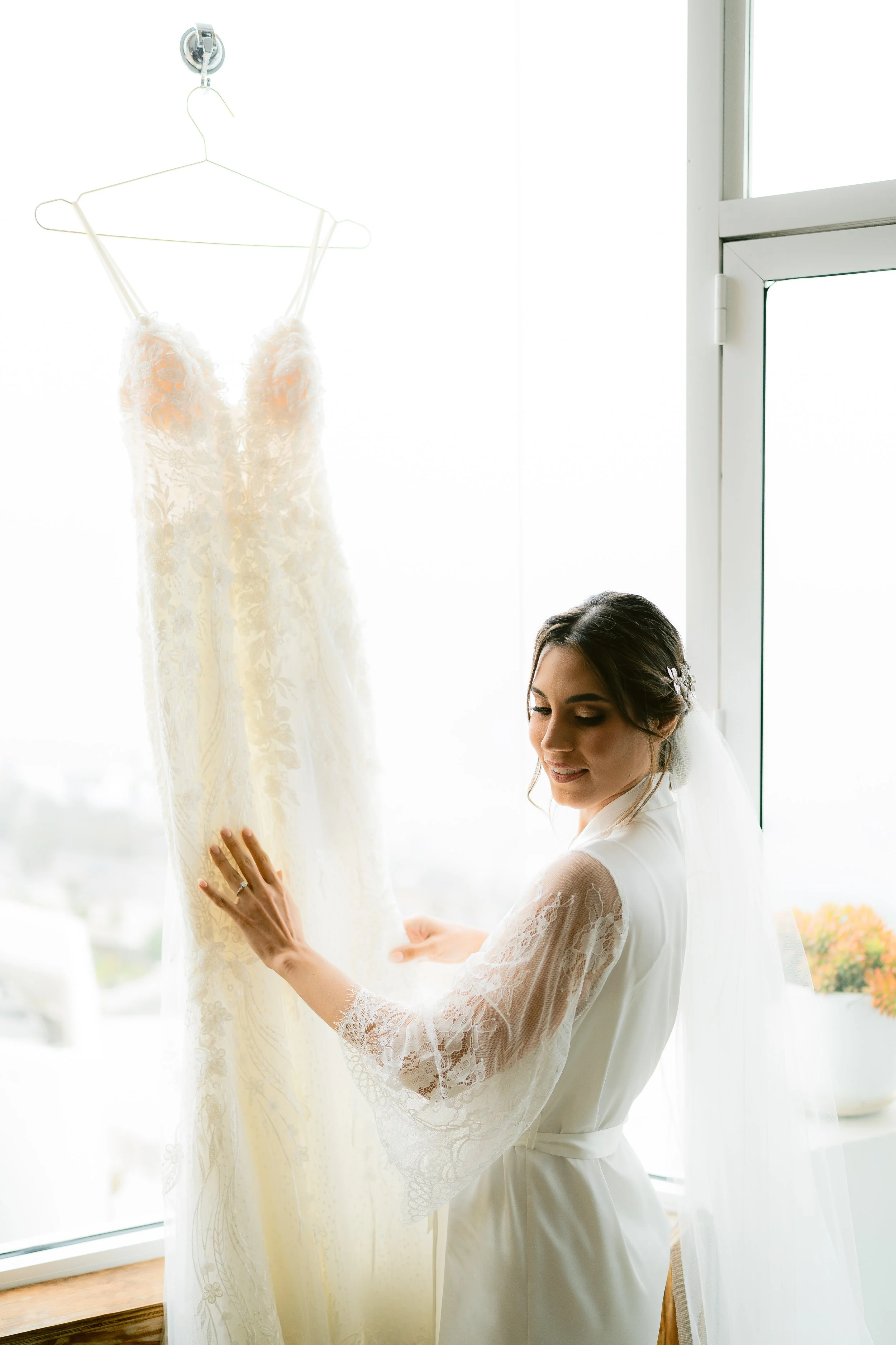 Una novia mira un vestido de novia colgado en una percha junto a una ventana.