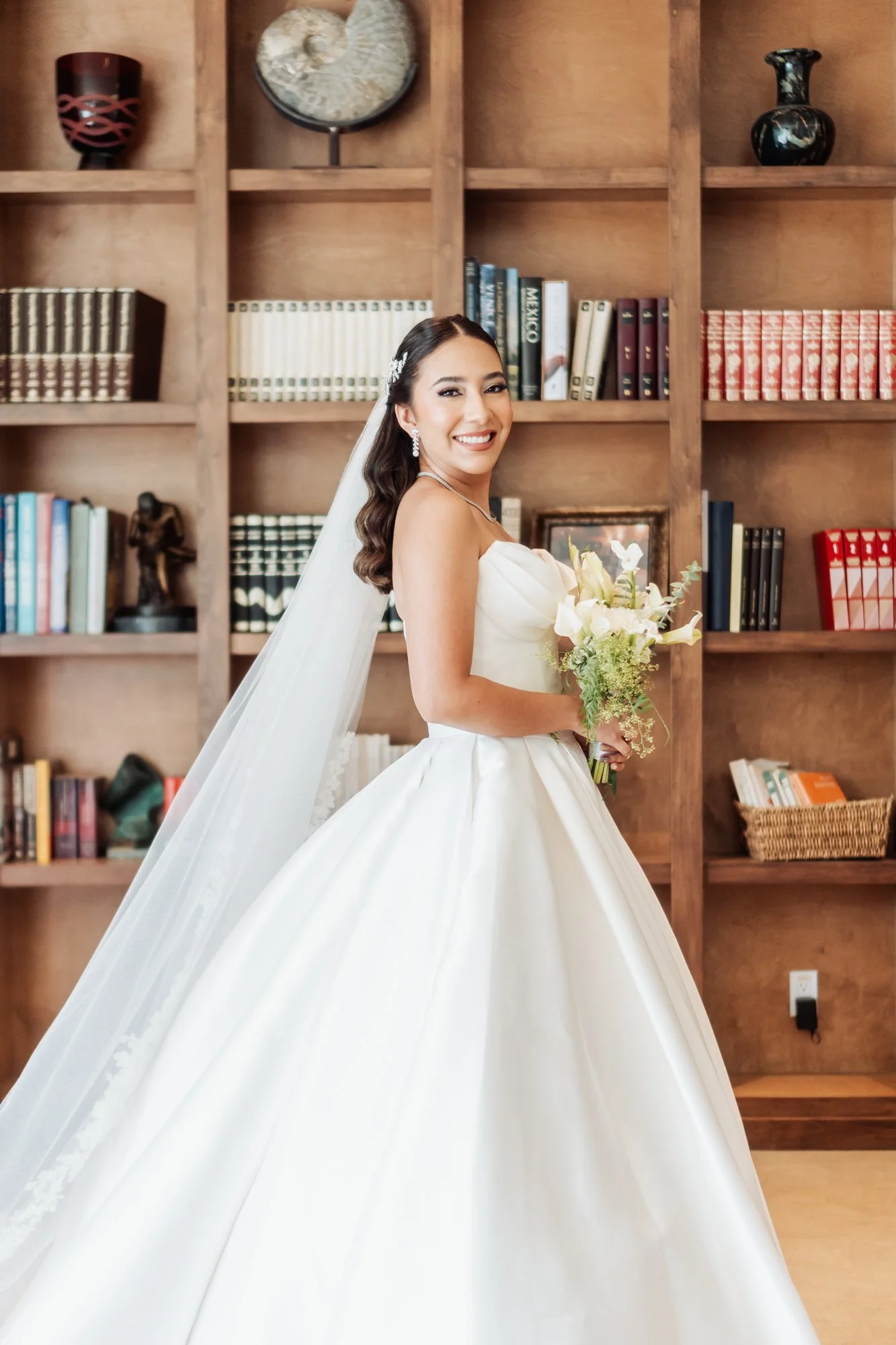 Mujer en vestido de novia sosteniendo un ramo de flores, sonriendo, con estantería de libros y decoraciones en el fondo.