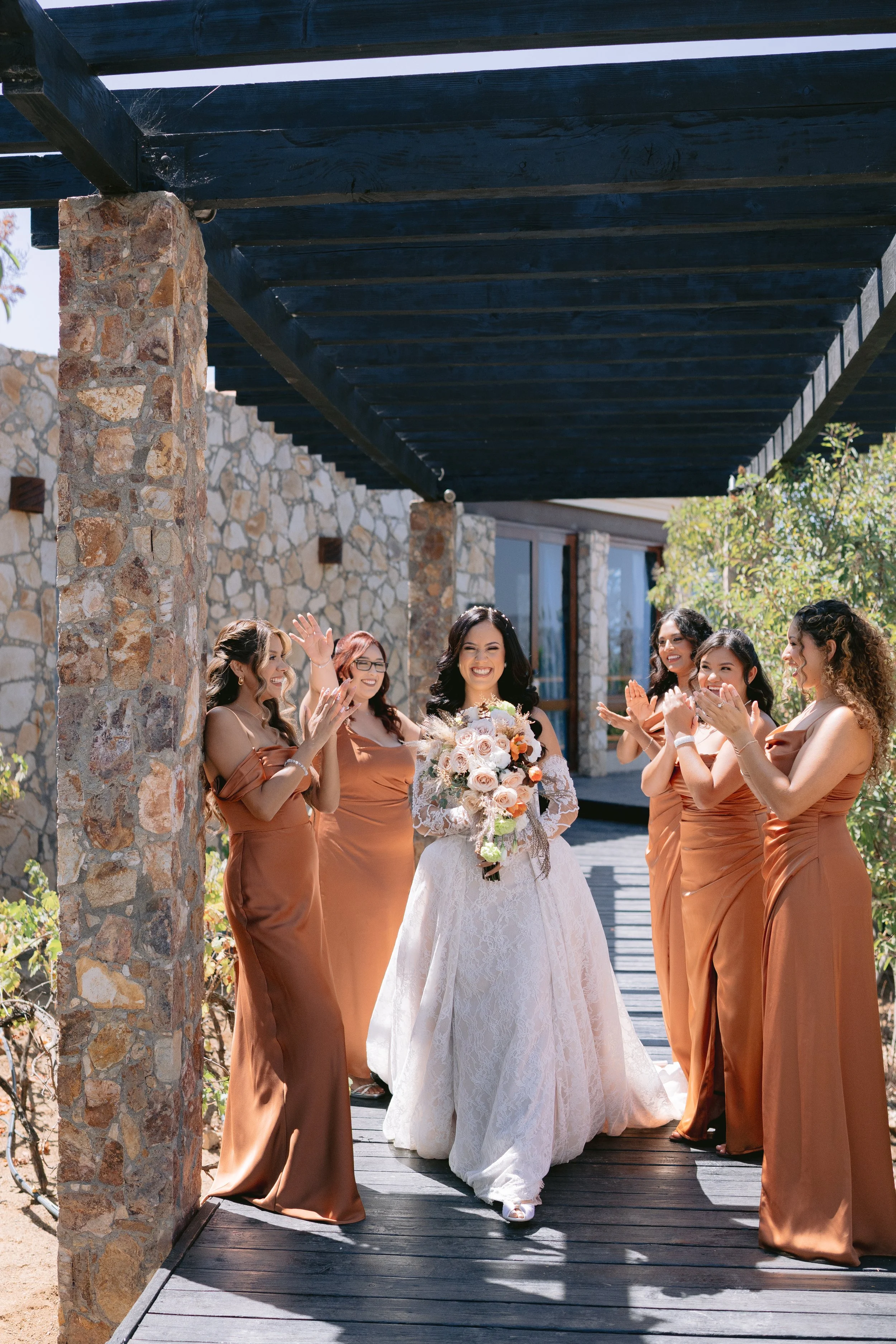 Mujer con vestido de novia sosteniendo un ramo de flores, rodeada de damas de honor en vestidos color bronce, en una ceremonia de boda al aire libre con estructura de madera y paredes de piedra.