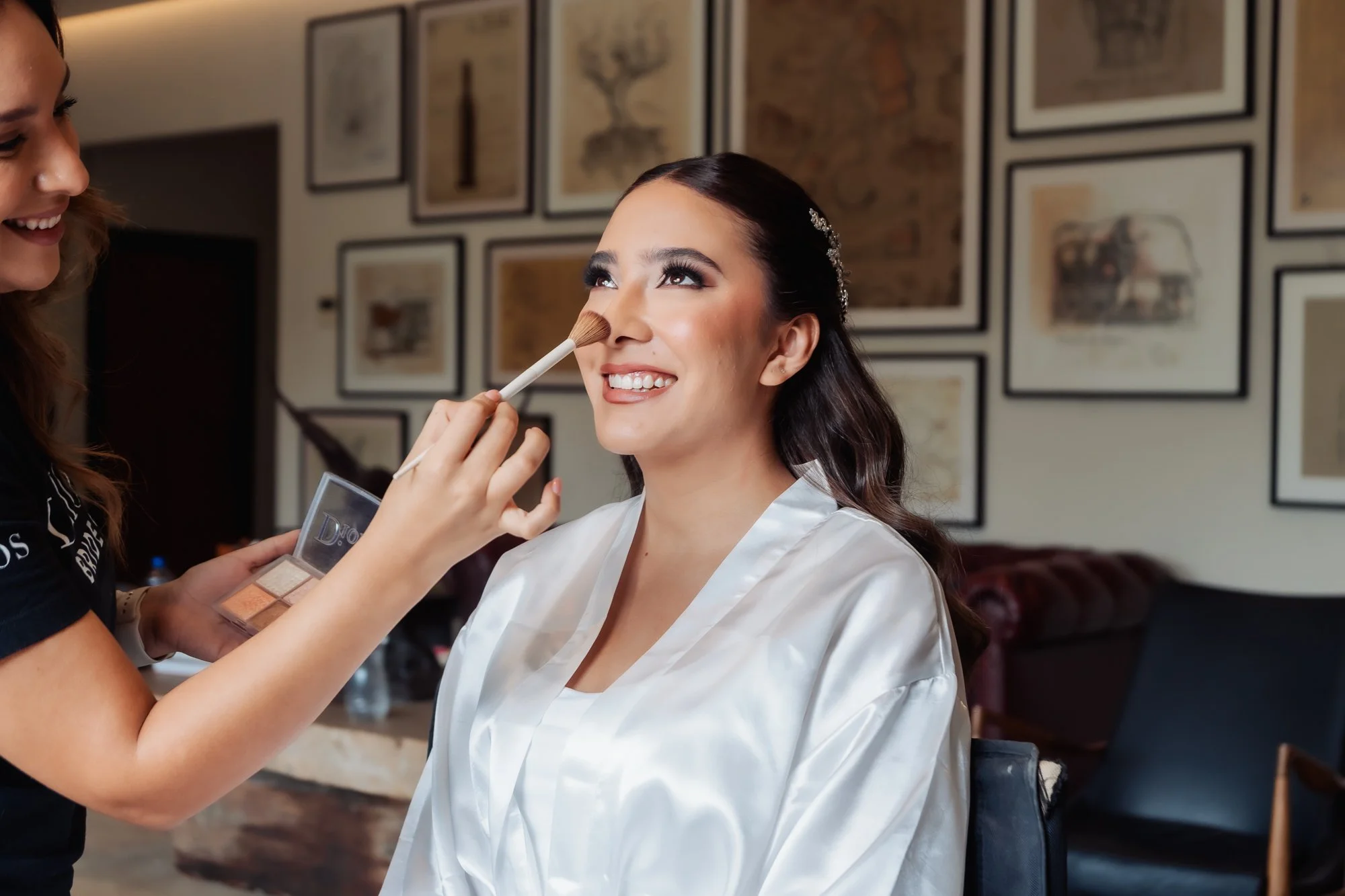 Una mujer con bata de seda blanca recibe ayuda para maquillar su rostro en un salón decorado con cuadros en la pared.
