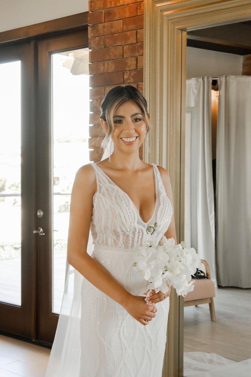 Mujer en vestido de novia sosteniendo un ramo de flores blancas en un interior con paredes de ladrillo y puertas de cristal.