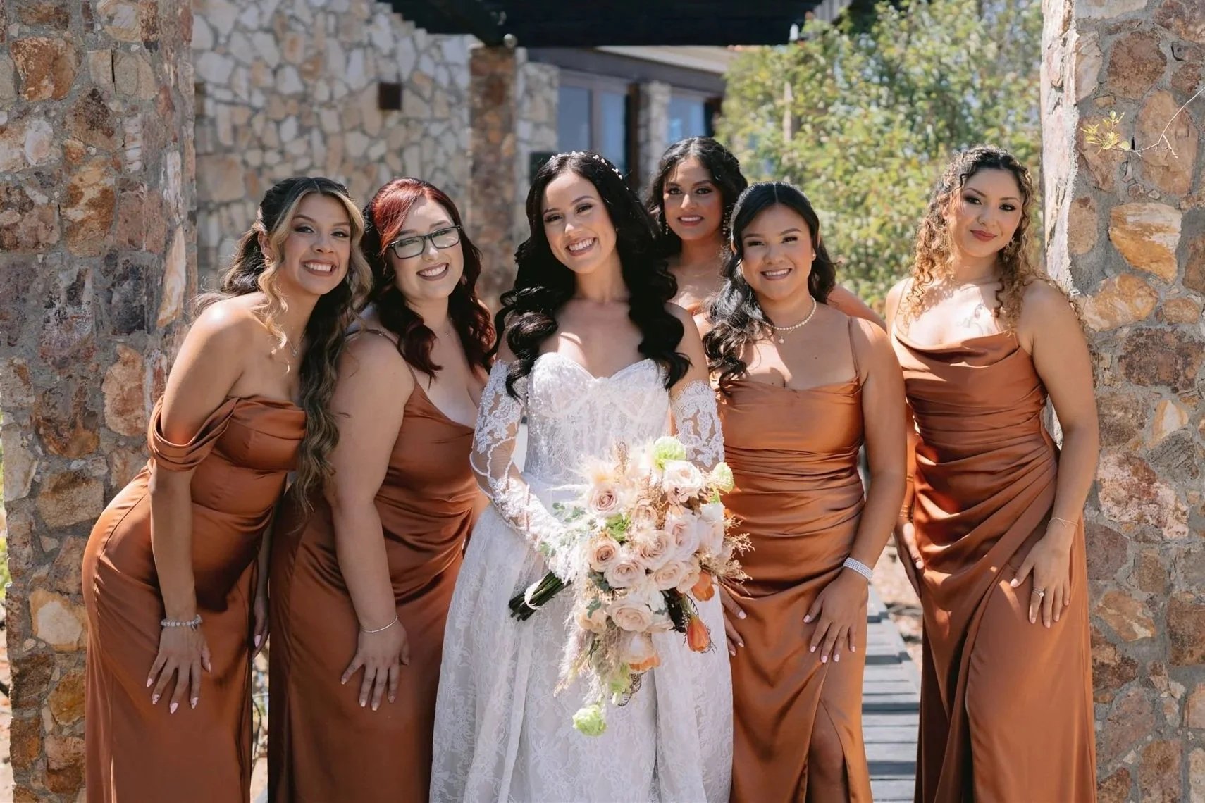 Grupo de mujer en vestido de novia y damas en vestido color canela posando junto a muro de piedra en un día soleado.