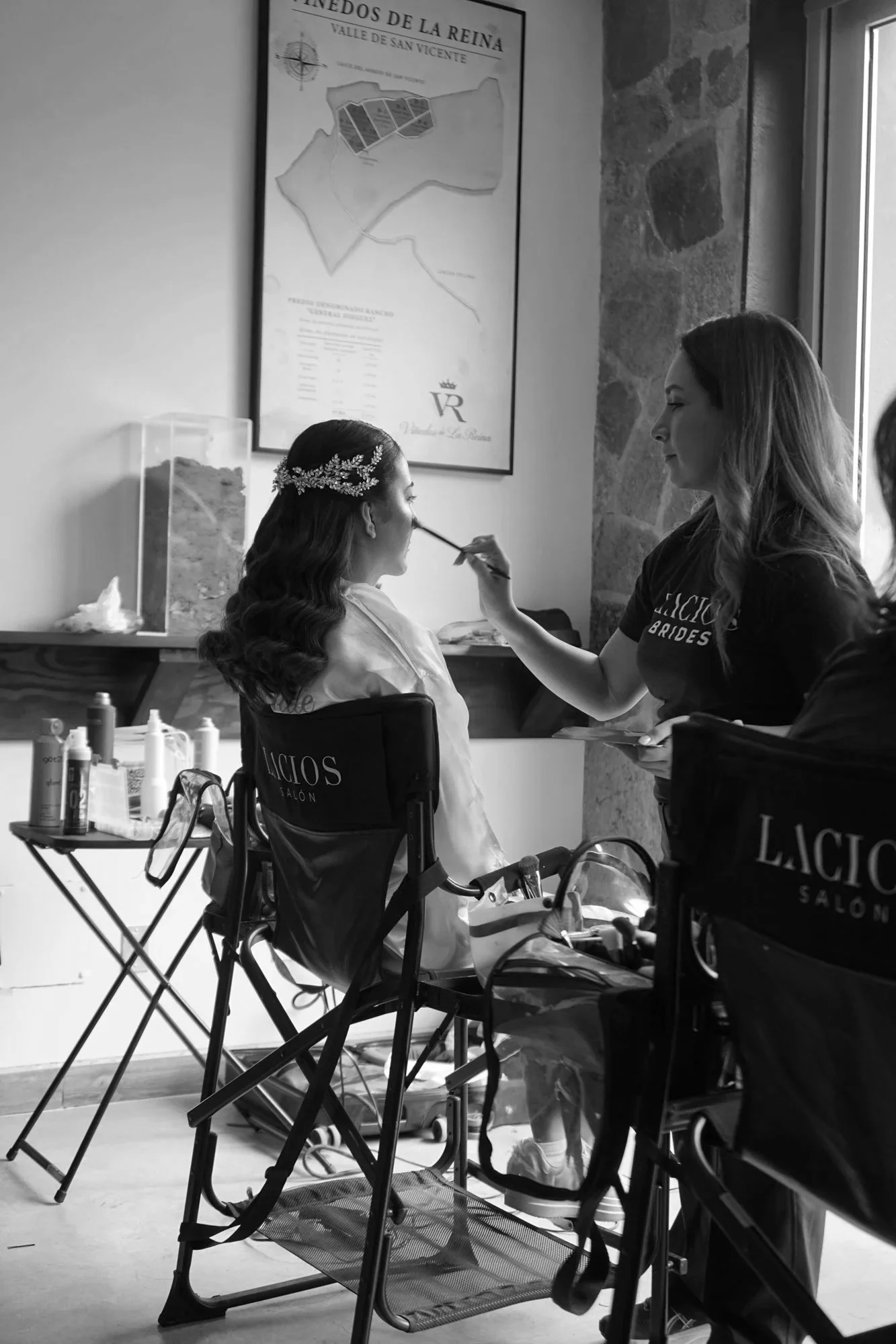 Mujer posando para maquillaje con otra mujer aplicándole maquillaje en un salón de belleza, con un mapa en la pared y materiales de maquillaje sobre una mesa.