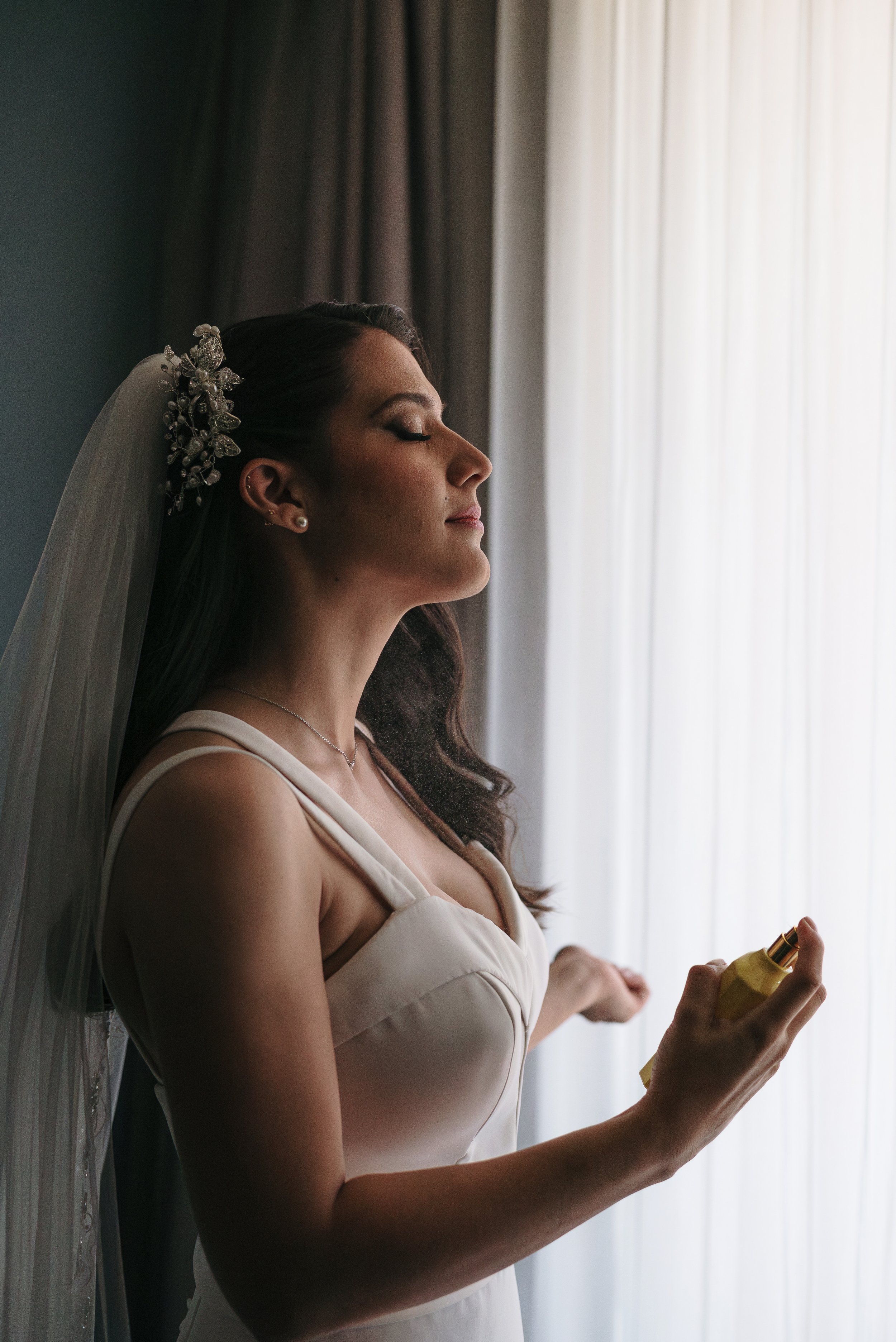 Mujer con vestido blanco y velo, con peinado y accesorios elegantes, aplicando perfume en una habitación con cortinas blancas.