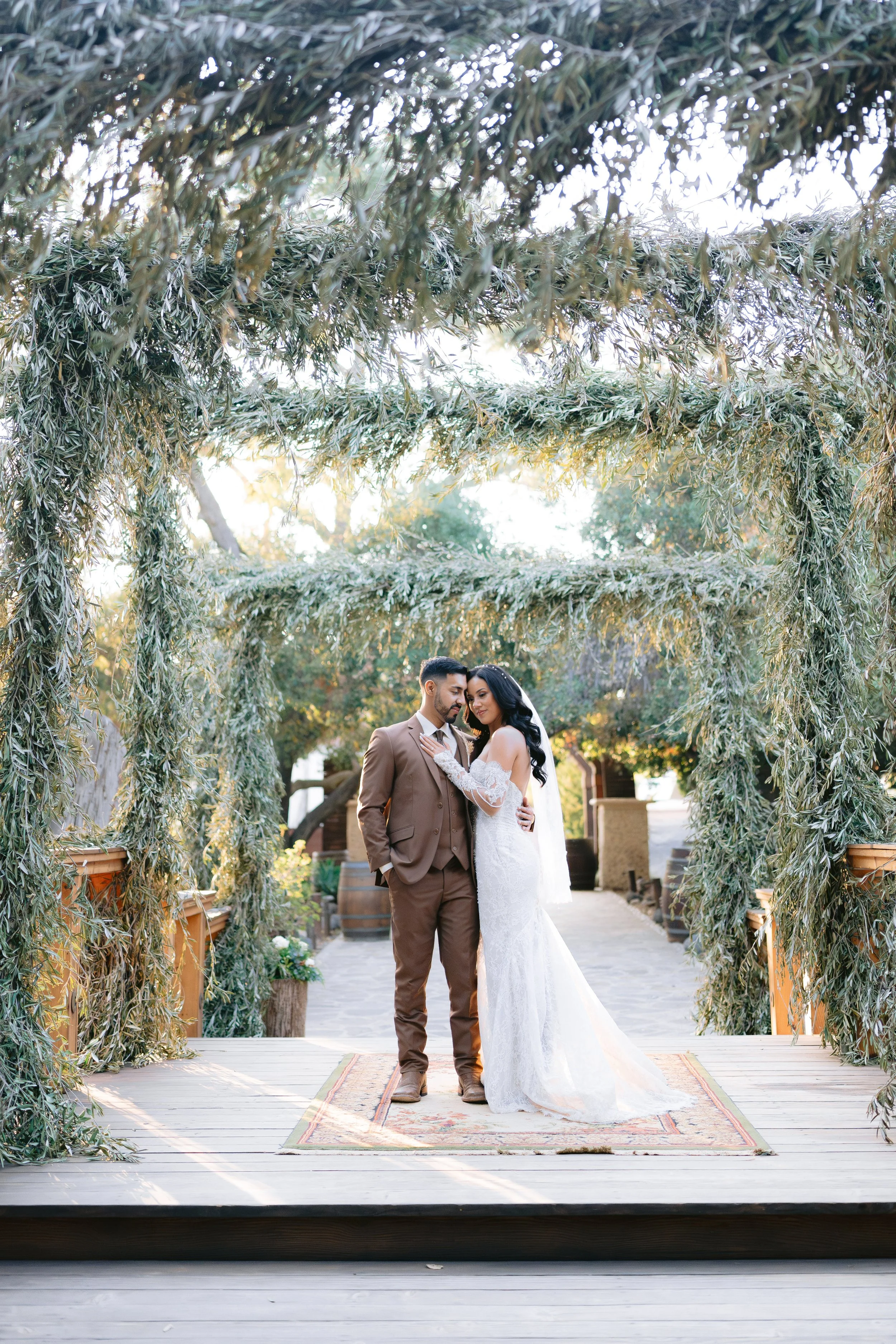Una pareja de novios en vestimenta nupcial en una ceremonia al aire libre, frente a un arco decorado con ramas verdes y plantas, en un entorno natural y soleado.