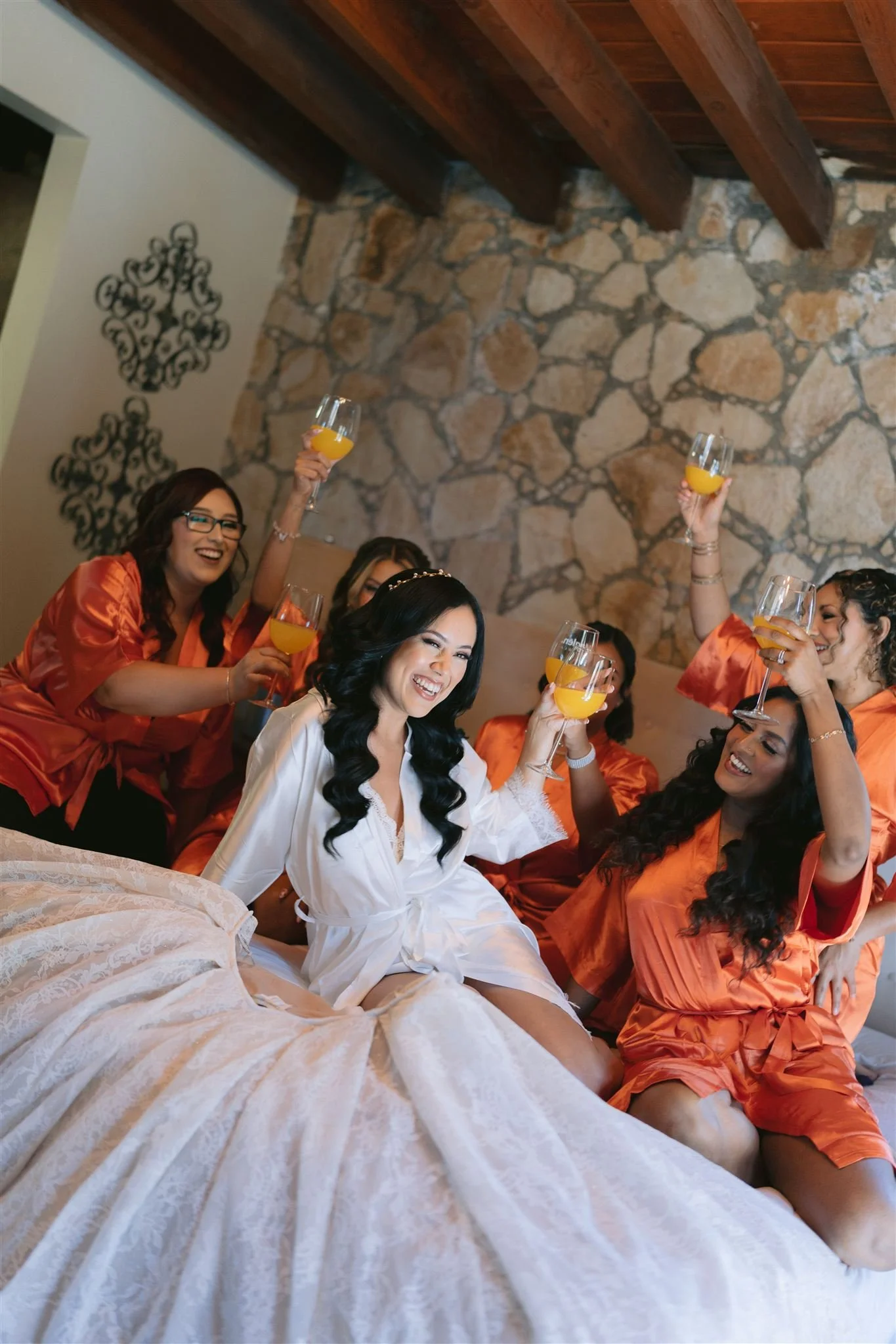 Grupo de mujeres en pijama celebrando con copas de jugo de naranja, en una habitación con paredes de piedra y decoración floral, probablemente en preparación para una boda o evento especial.