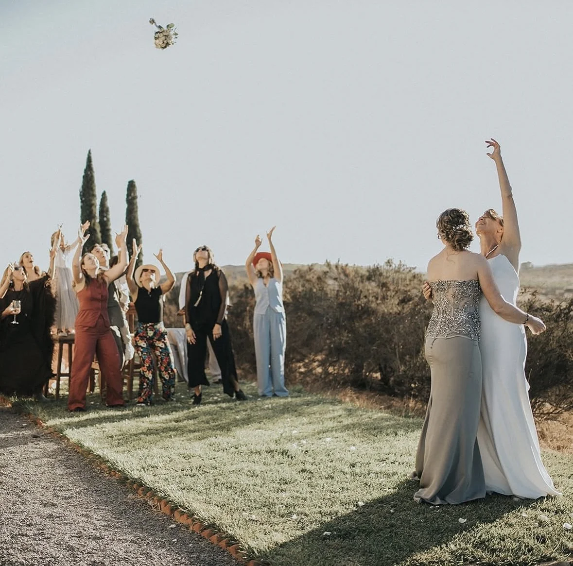 Pareja en vestido de novia bailando en una boda al aire libre, con invitados en el fondo lanzando un ramo.