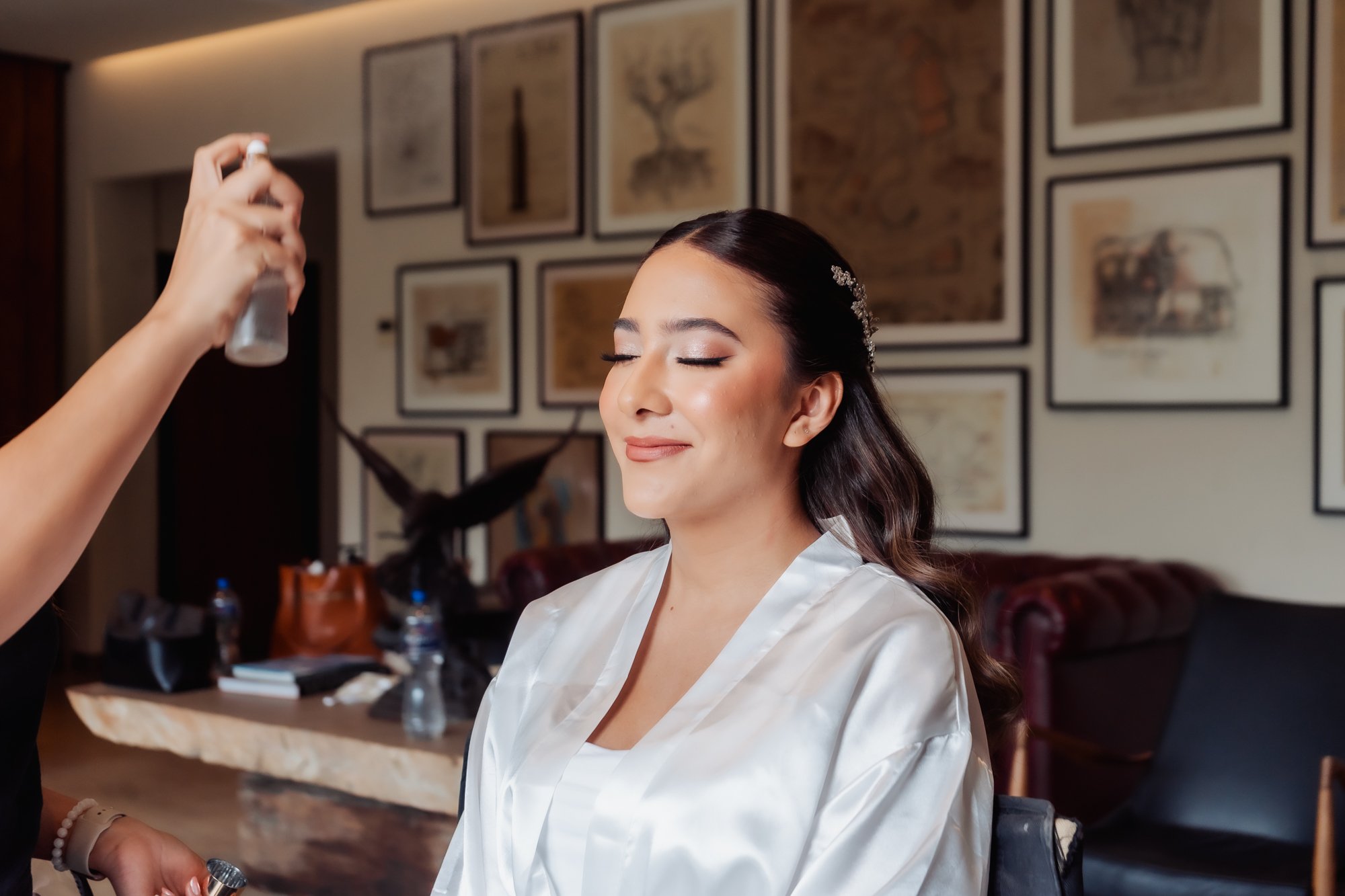 Mujer sonriendo con ojos cerrados mientras recibe un spray en la cara, vestida con bata de satín blanca.