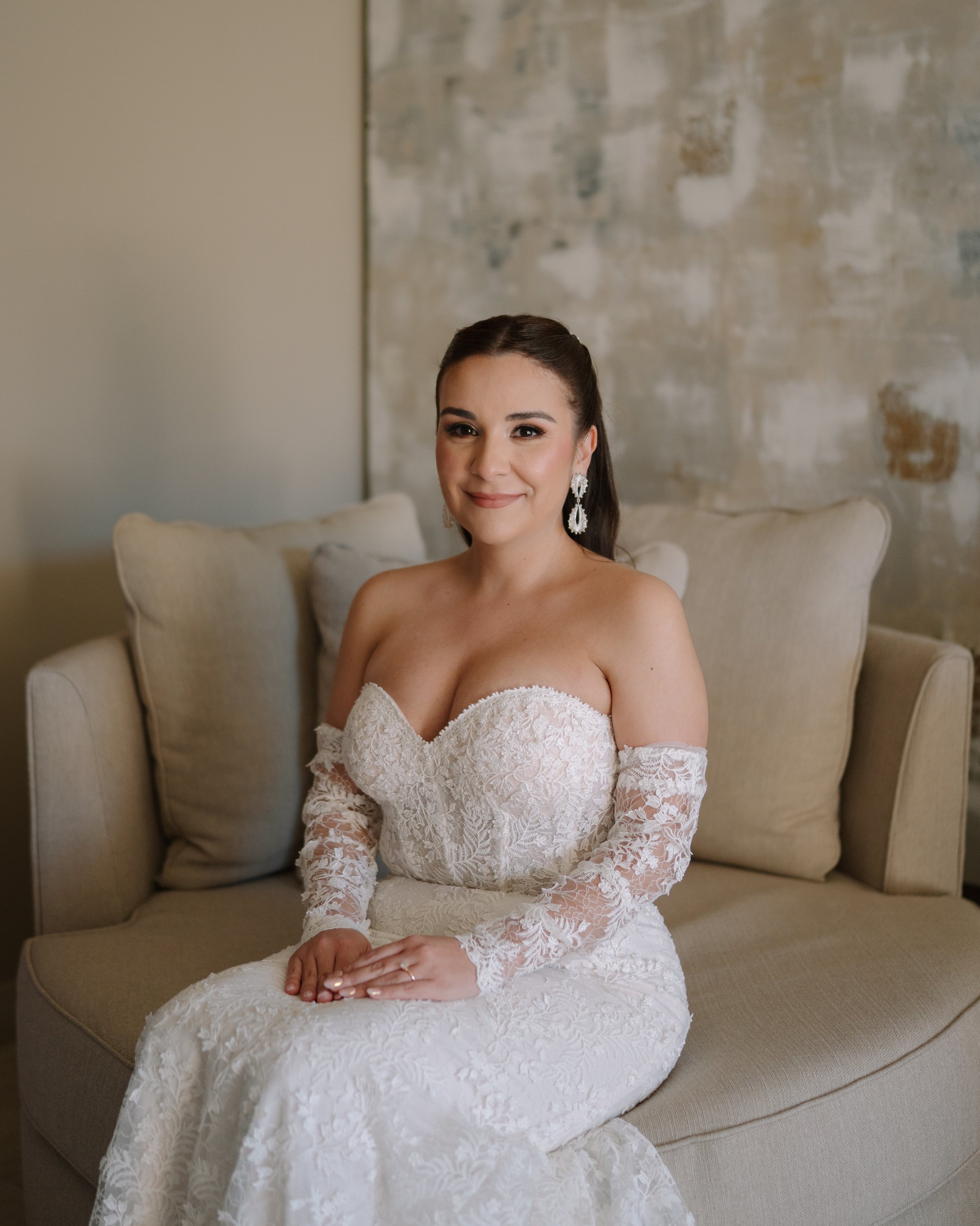 Mujer en vestido de novia sentado en un sillón, sonriendo, con fondo de pared de colores neutros.