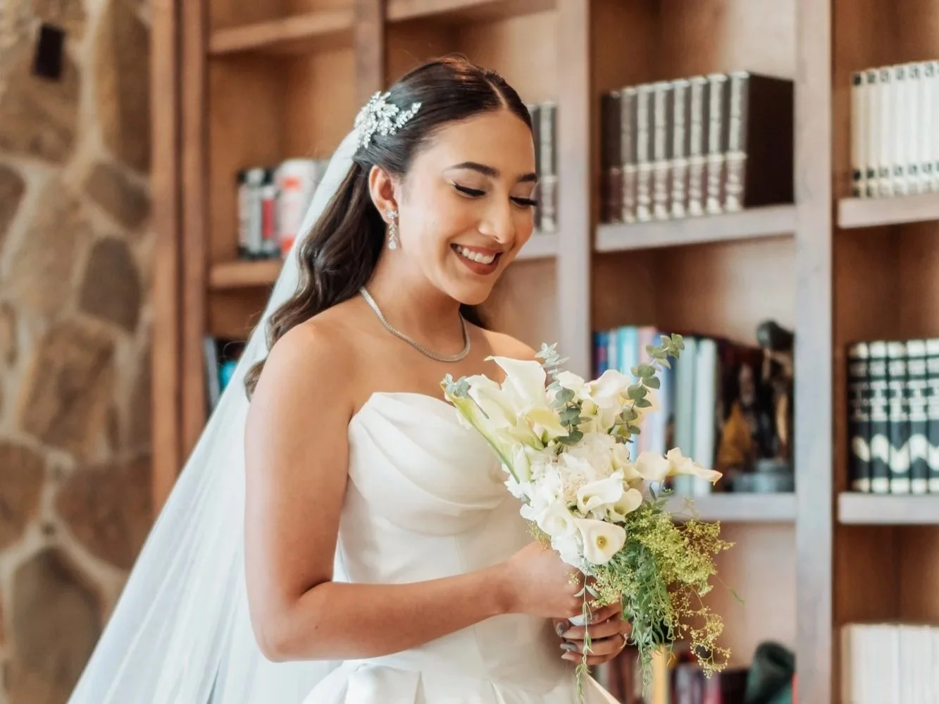 Mujer vestida de novia en una habitación con libreros de madera, sosteniendo un ramo de flores blancas.