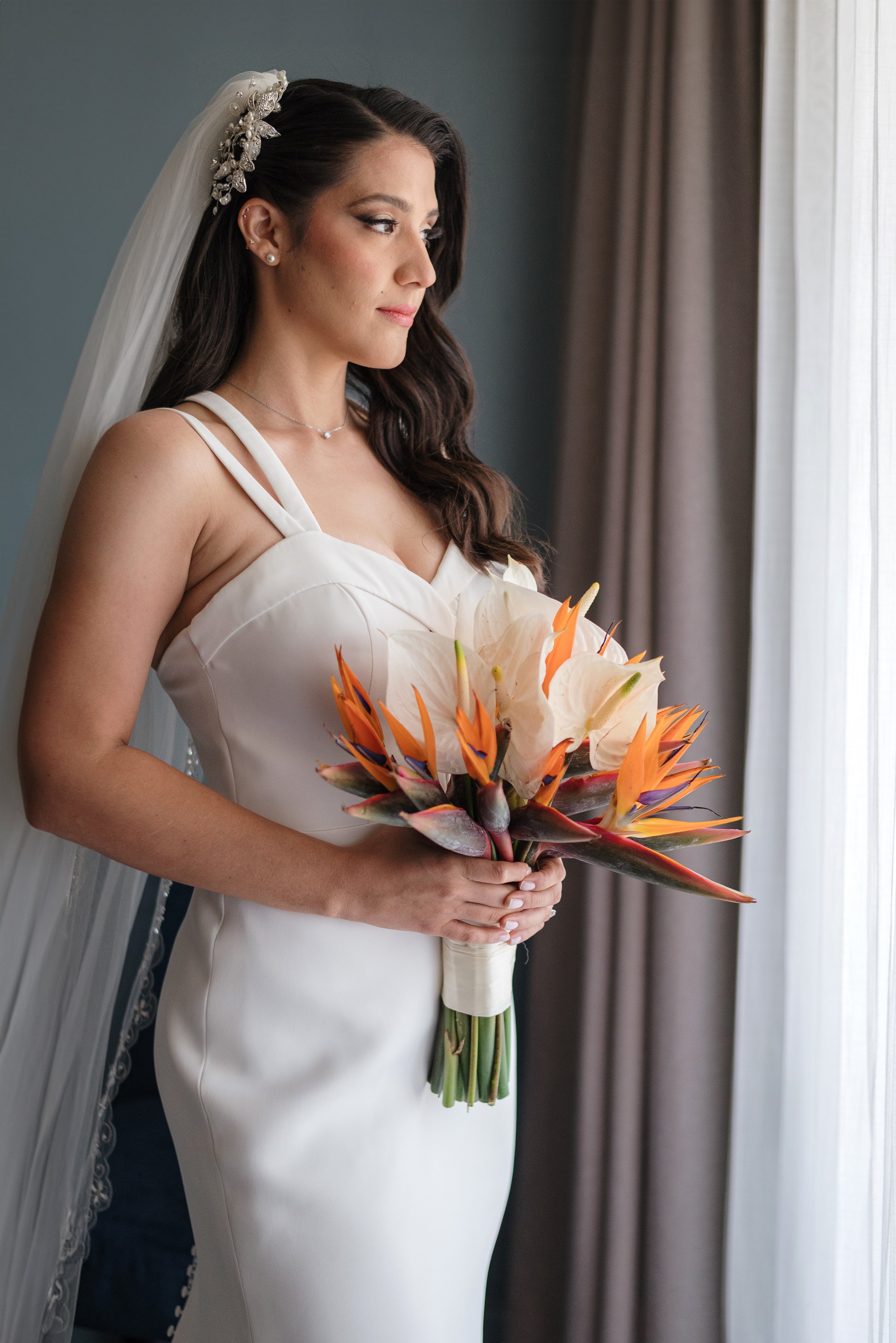 Mujer en vestido de novia con ramo de flores blancas y coloridas, mirando por la ventana