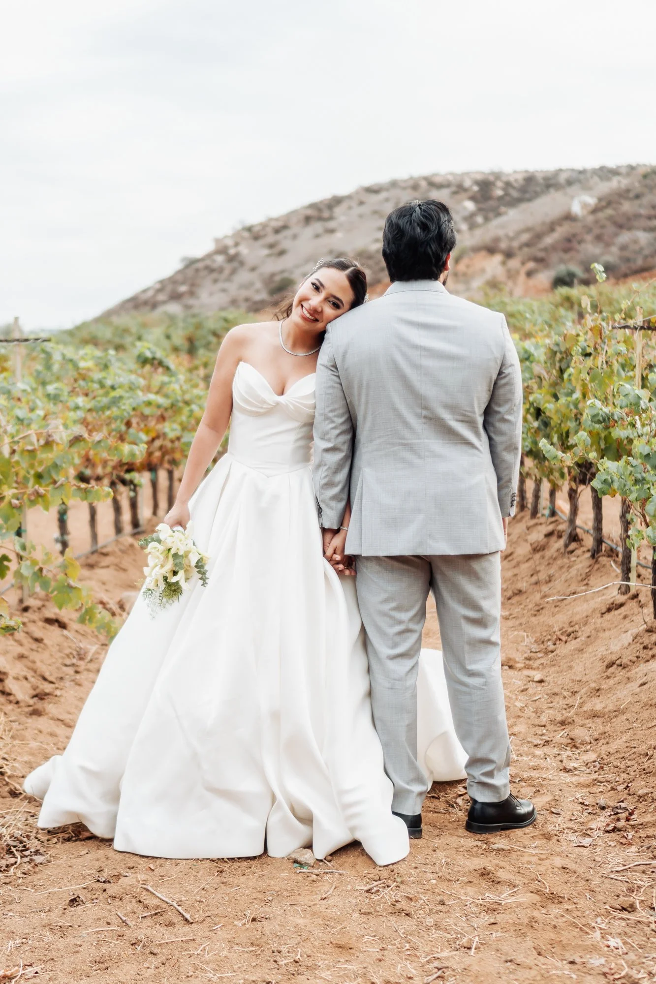 Una pareja de recién casados en un viñedo, la mujer sonríe y apoya su cabeza en el hombro del hombre, que está de espaldas, ambos con ropa de boda.