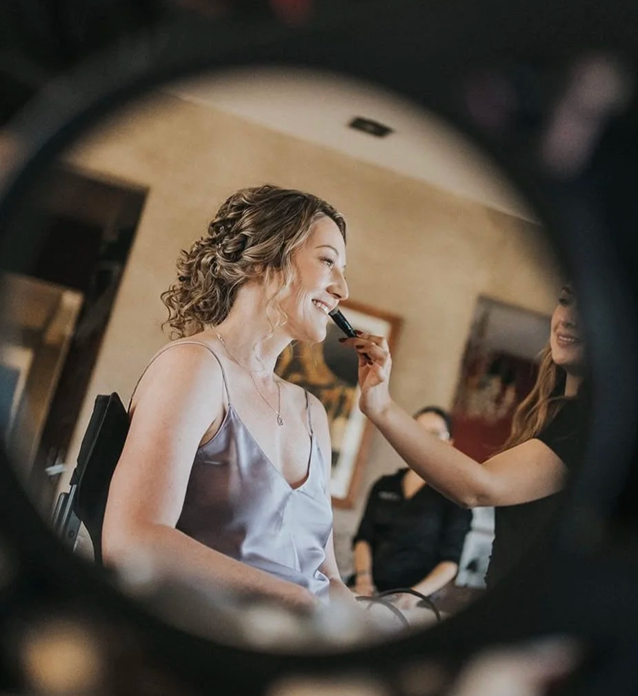 Mujer joven con cabello rizado y vestido gris sentada, recibiendo maquillaje o peinado por otra mujer, en un entorno interior con cuadros en la pared.
