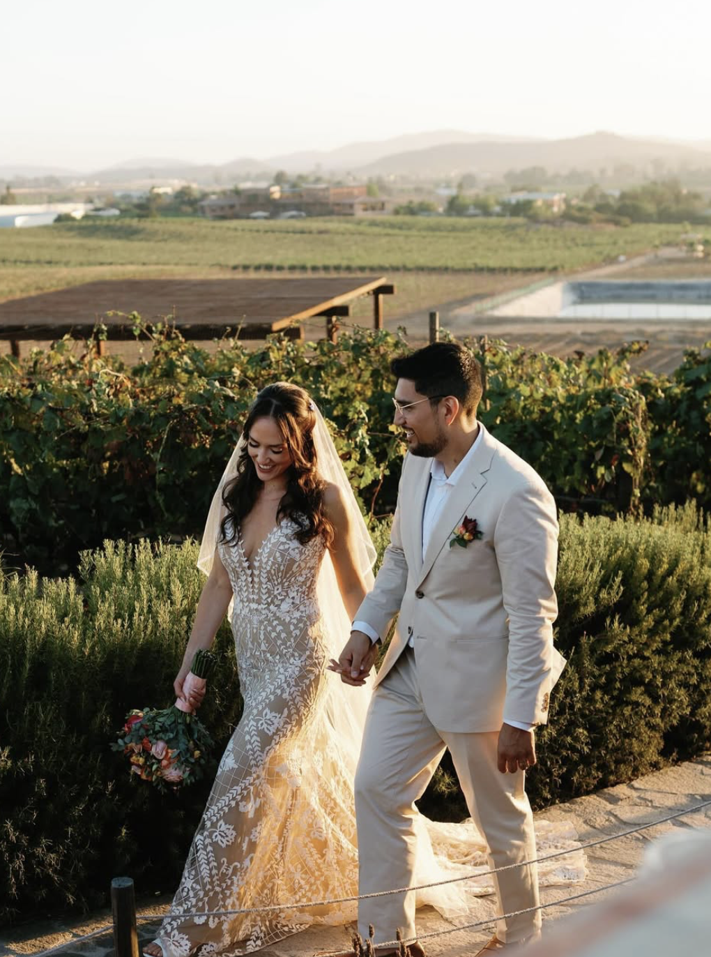 Una pareja de boda caminando en un viñedo al atardecer, la mujer lleva un vestido de encaje y el hombre usa un traje crema.