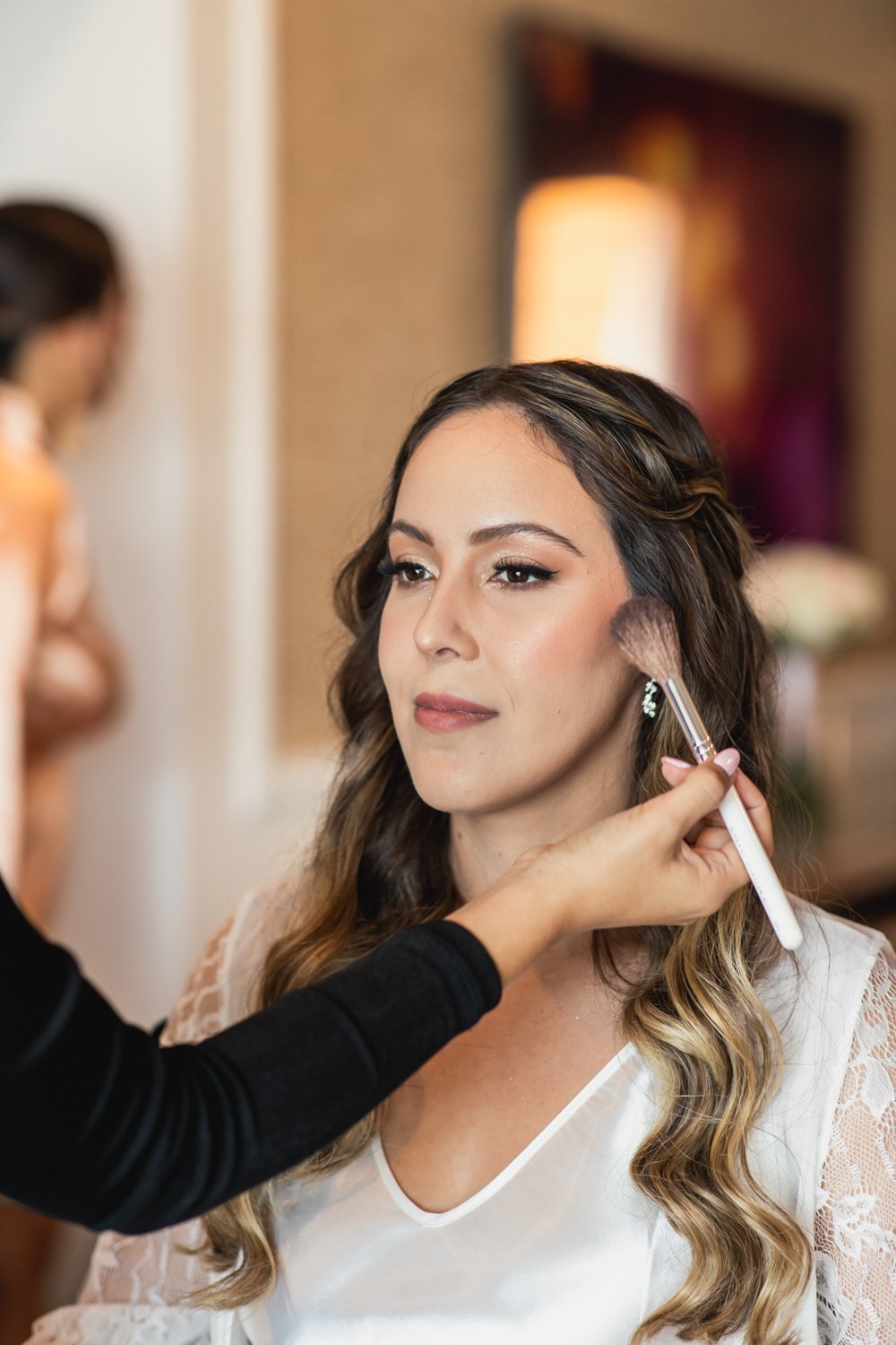 Mujer con cabello castaño y rubio recibiendo los últimos retoques de maquillaje por parte de una maquilladora en un ambiente interior, probablemente antes de un evento especial.