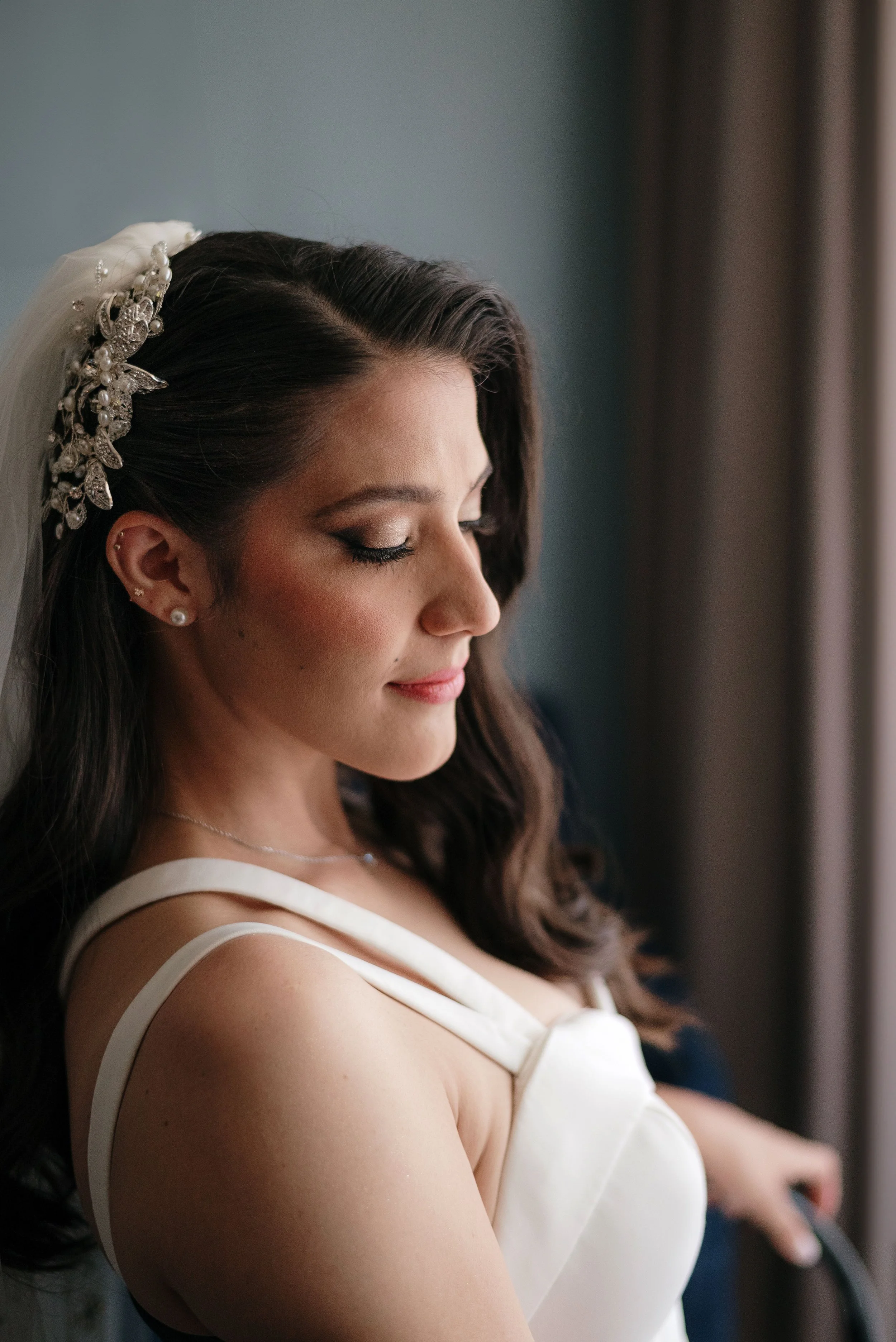 Joven novia con cabello castaño oscuro, peinada con accesorios florales y cristales, usando vestido de novia blanco, mirando hacia abajo con ojos cerrados, en un ambiente con luz natural.