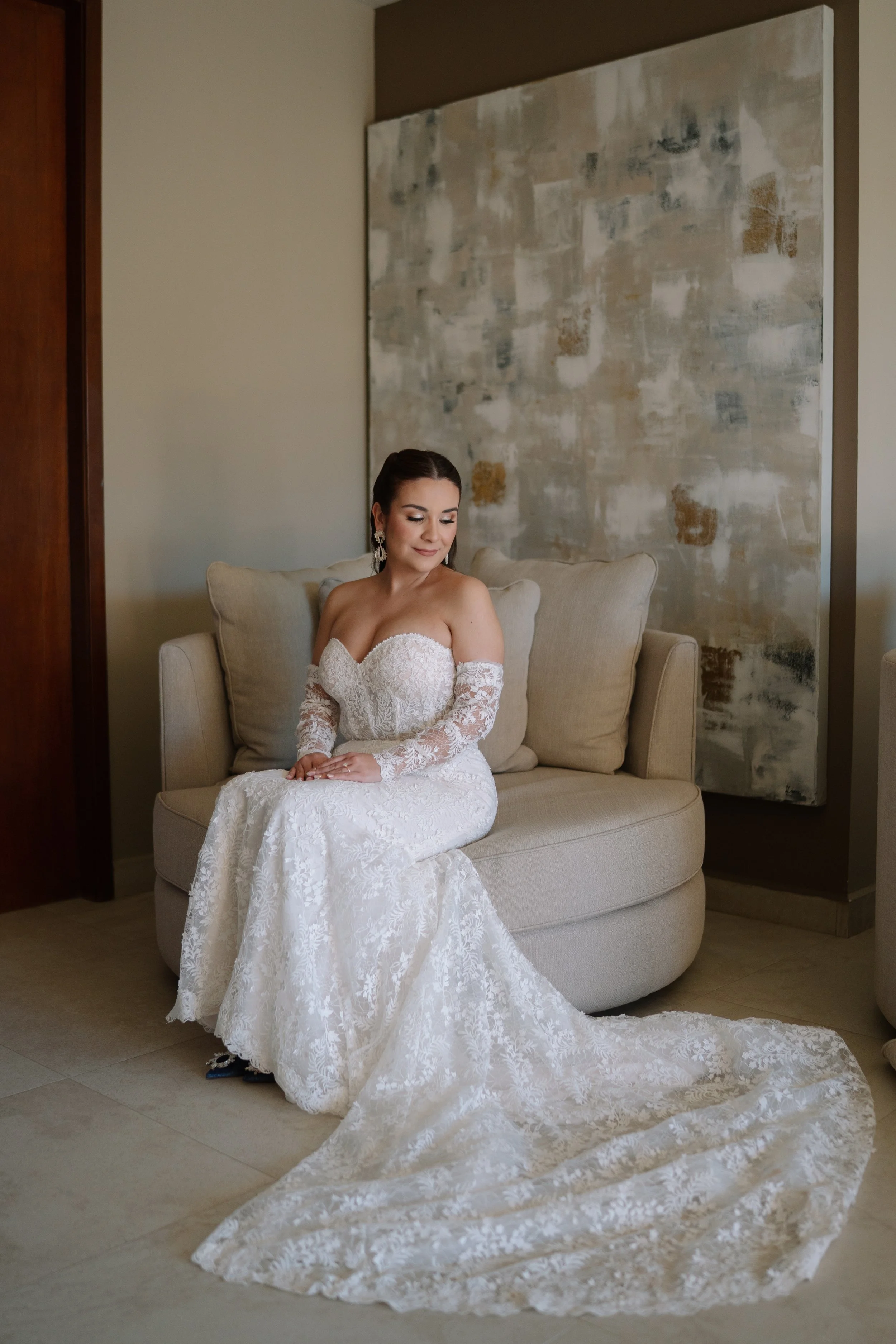 Mujer en vestido de novia sentado en un sofá beige en un interior decorado con cuadros abstractos.  maquillista de novias en ensenada y valle de guadalupe 