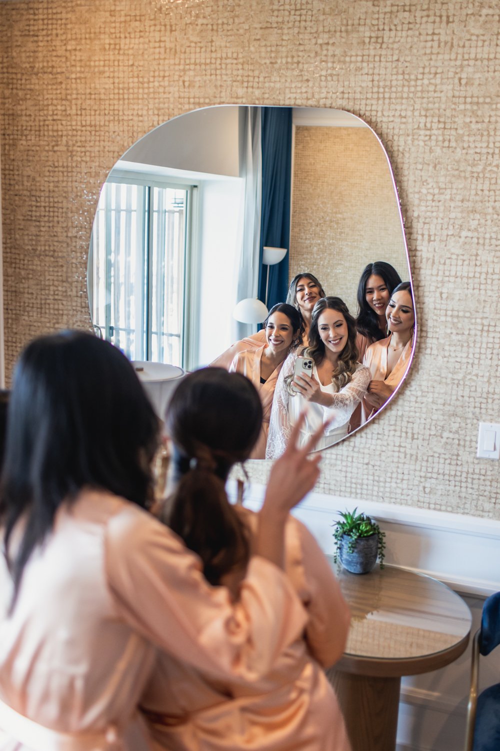 Grupo de mujeres sonriendo y tomándose una selfie frente a un espejo en una habitación bien iluminada.
