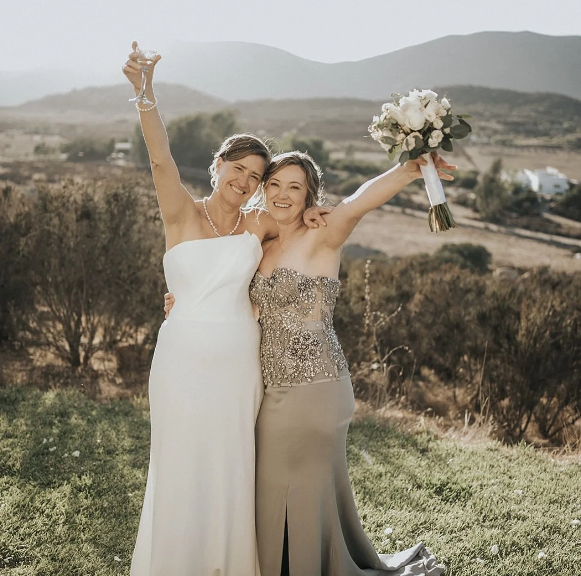 Dos mujeres celebrando en un campo abierto, una con vestido de novia y la otra con vestido de fiesta, ambas sonriendo y levantando los brazos, una con un ramo de flores blancas. Parece ser una celebración especial.