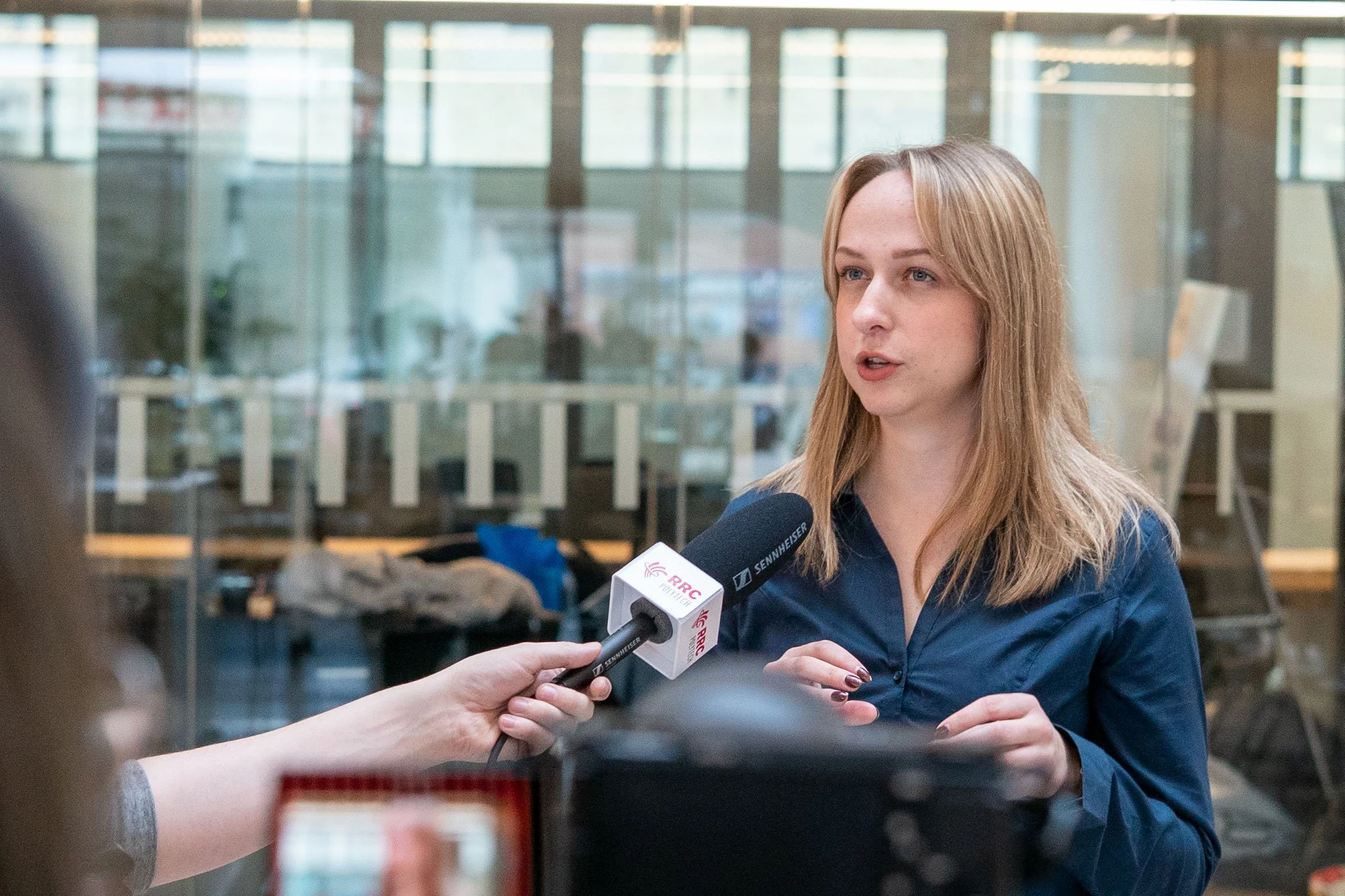 A woman with red hair wearing a navy blue shirt is being interviewed indoors with a microphone labeled 'RRC' and 'Sennheiser' held near her mouth, with a glass wall and blurred figures in the background.
