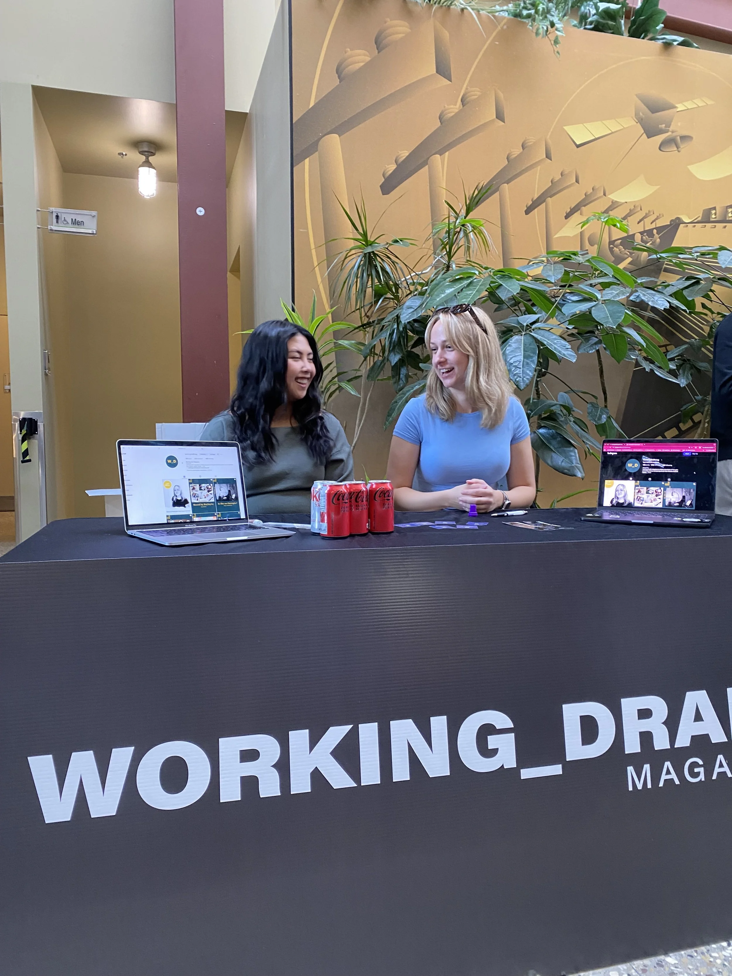 Two women sitting behind a table with laptops and soda cans, smiling and talking, at a booth with a "WORKING_DRAFT MAGAZINE" banner, in an indoor space with large green plants and wall art in the background.