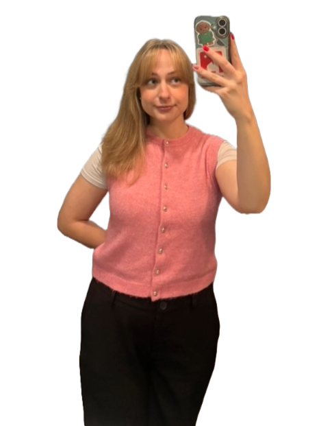 Young woman taking a mirror selfie with a smartphone, wearing a pink sleeveless cardigan over a white shirt and black pants.