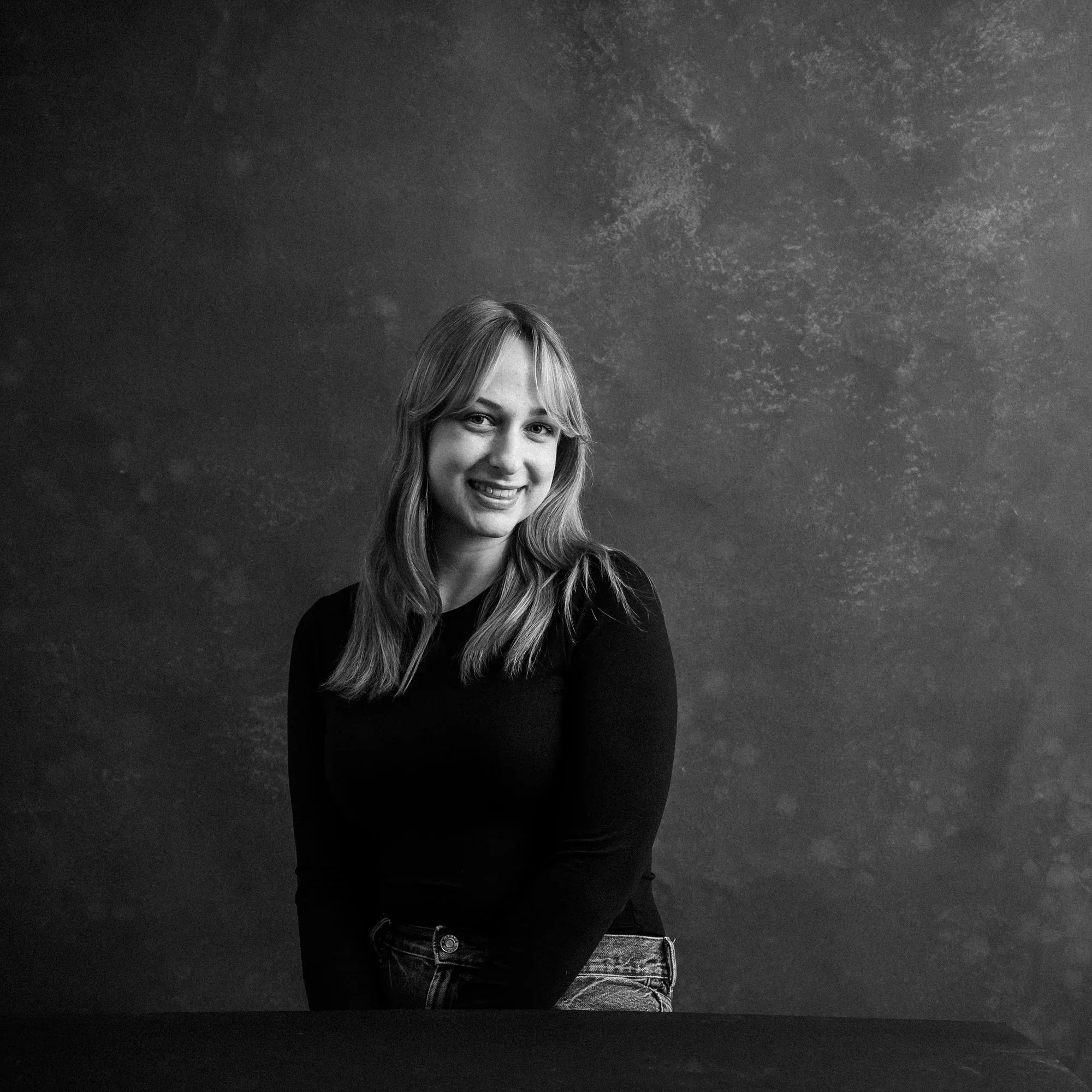 Black and white portrait of a young woman with long hair, smiling and wearing a black top and jeans, sitting against a textured background.