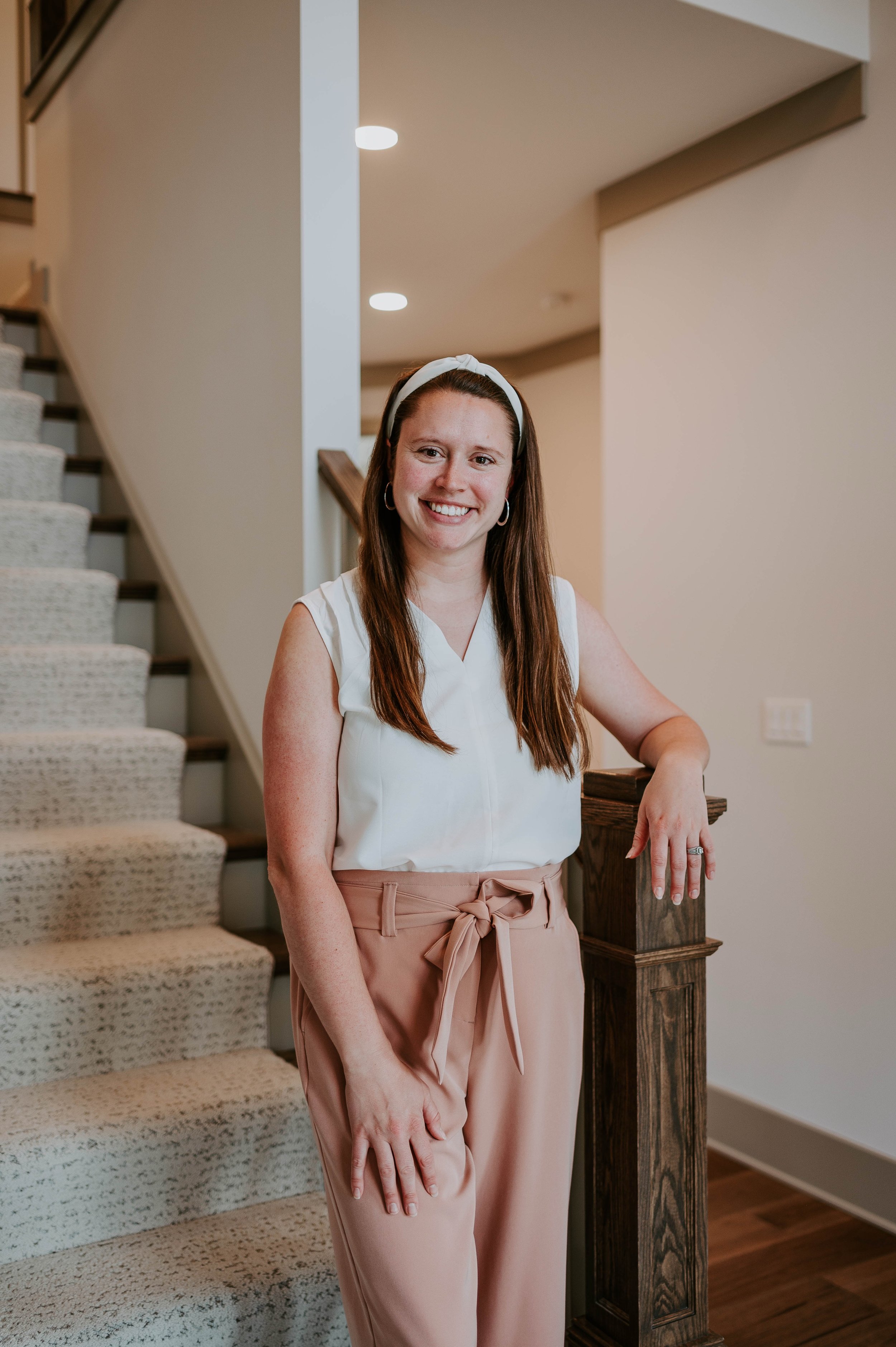 A woman with long brown hair wearing a white sleeveless top, pink high-waisted pants with a bow, and a white headband, standing indoors near a staircase with her hand resting on a wooden staircase post, smiling at the camera.