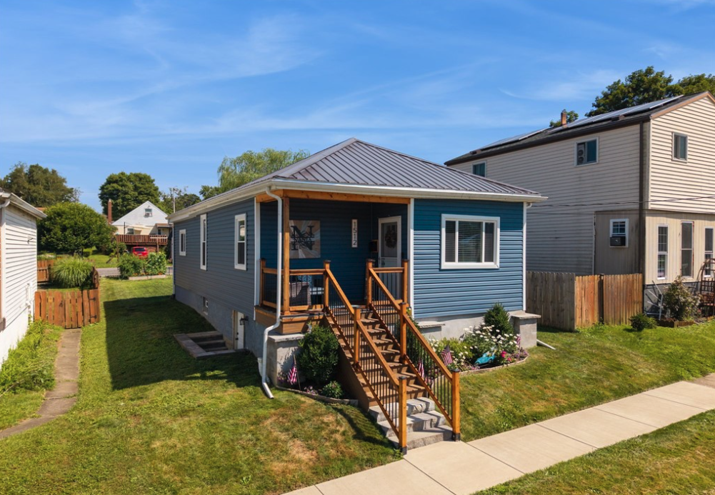 Blue house with wooden stairs and small front porch, surrounded by green lawn, flower garden, and neighboring houses under a blue sky.