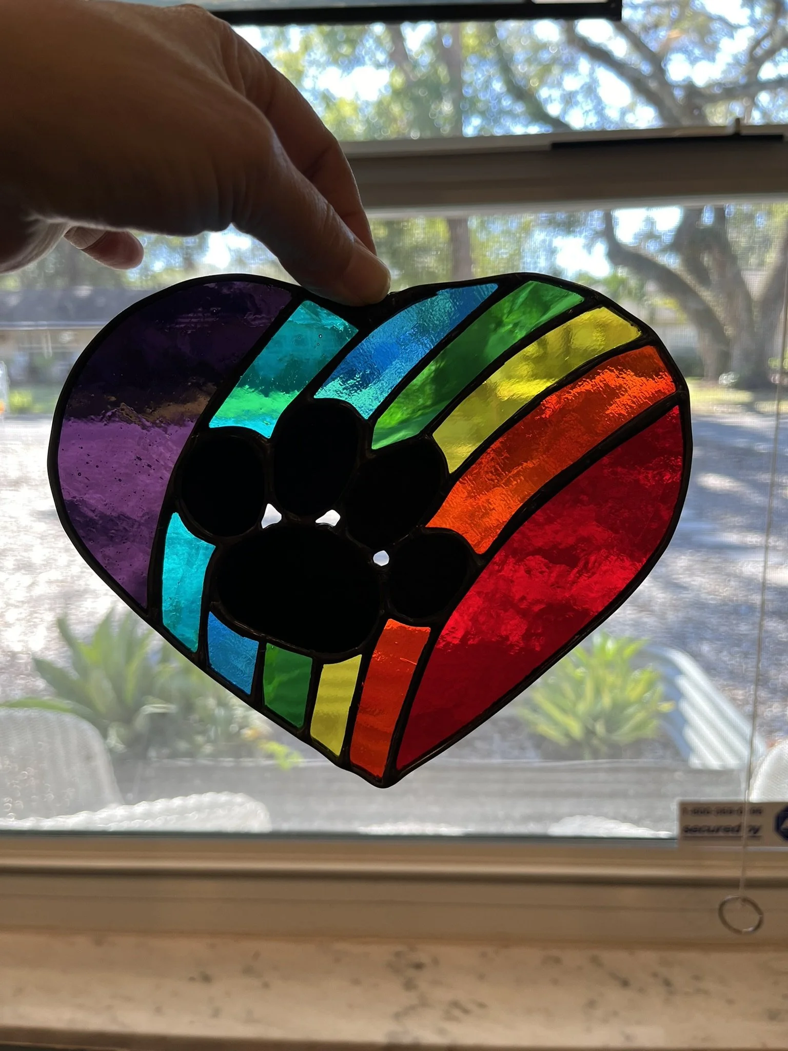 Colorful stained glass heart with a paw print design, held up in front of a window with a view of trees and outdoors.