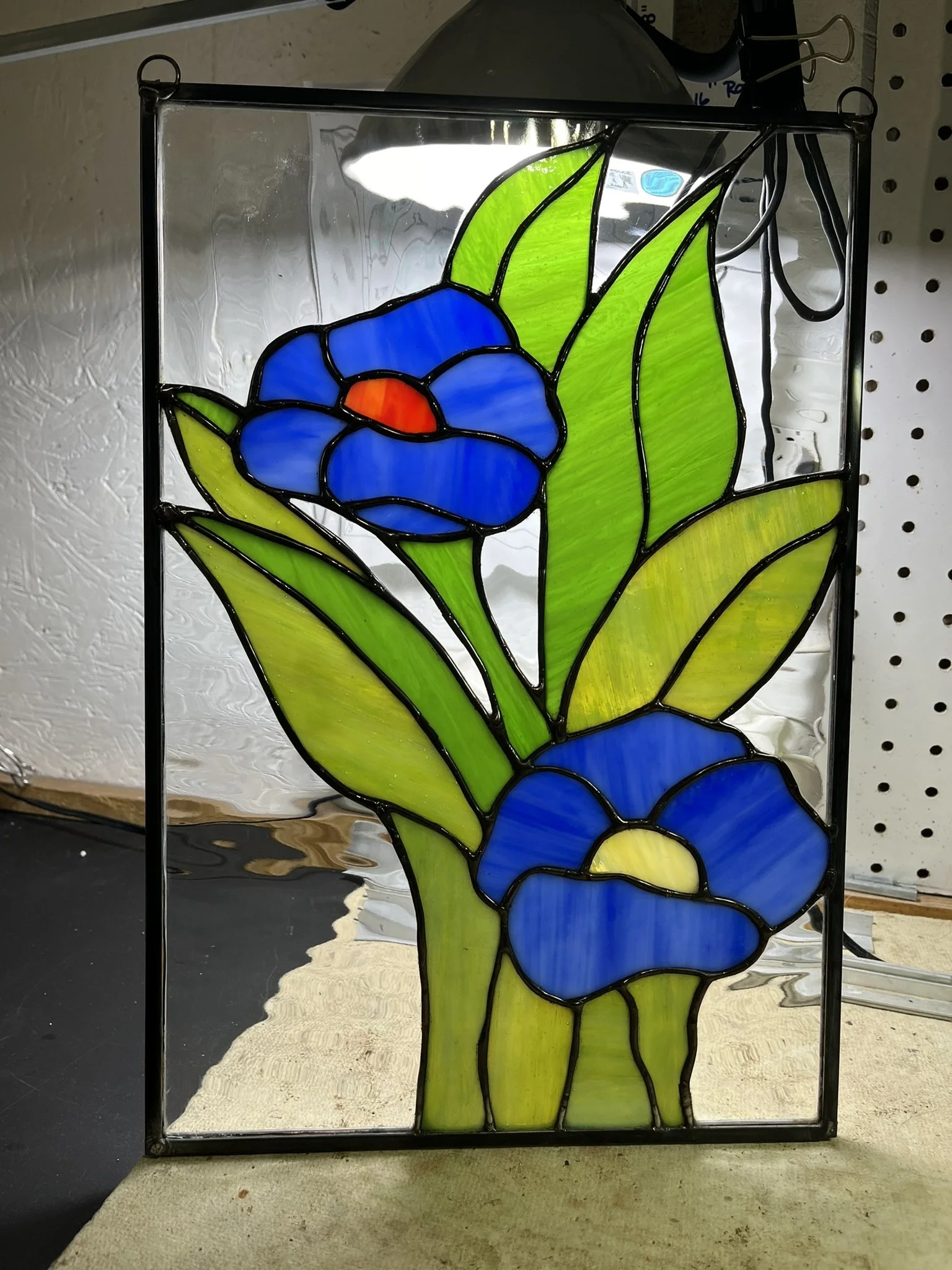 Stained glass artwork of two blue flowers with green leaves in a black frame.