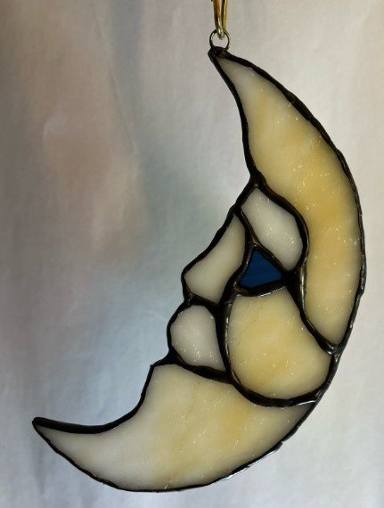 A stained glass sun and moon ornament with a crescent moon shape and stained glass segments in cream, blue, and black outlines, hanging from a metallic loop.