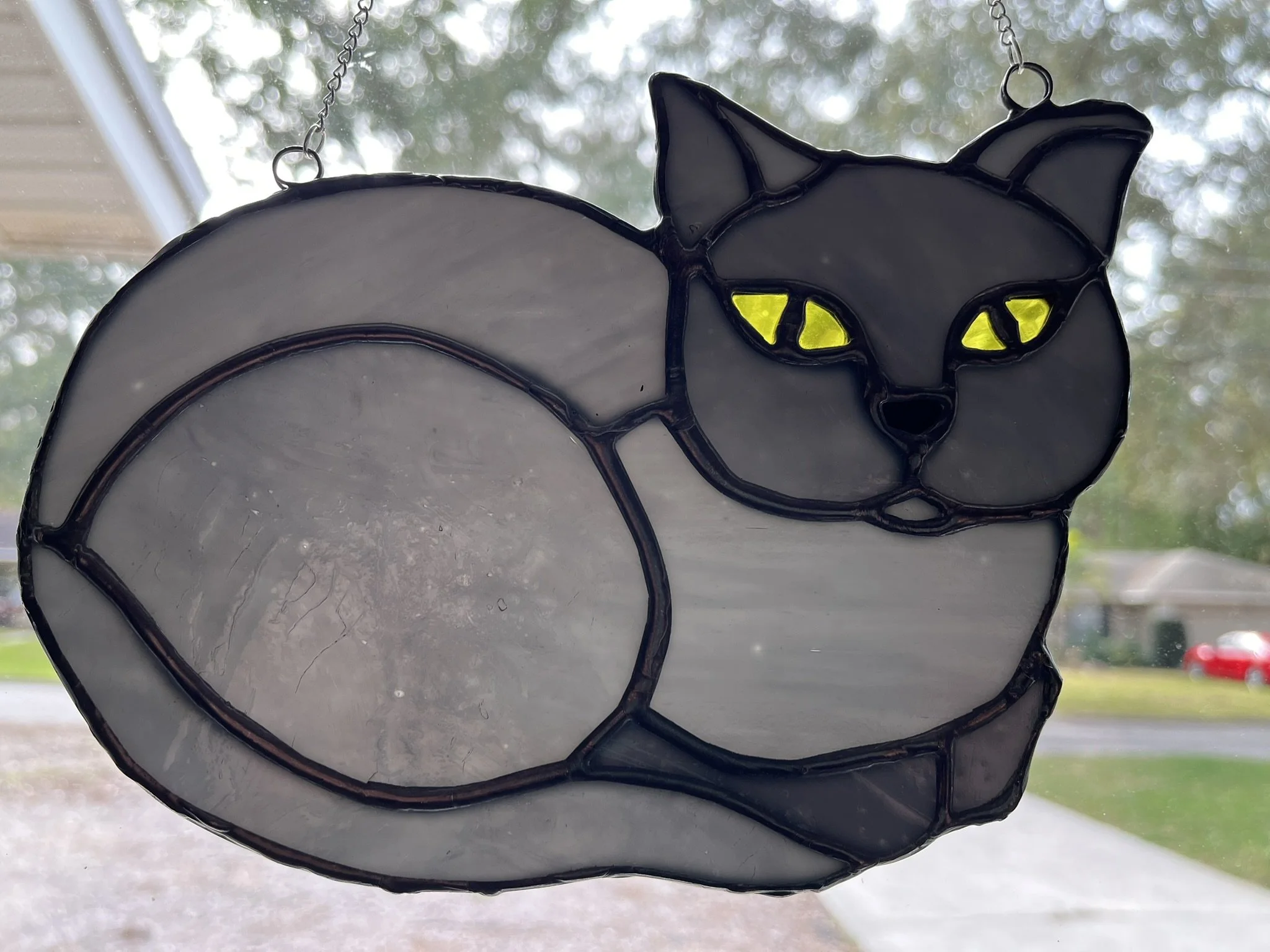 Stained glass sun catcher of a gray cat with glowing yellow eyes sitting with its tail curled around its body, hanging in front of a window.