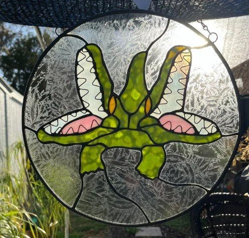 Seminole Heights Two Headed Gator stained glass window with green and yellow accents, hanging outdoors with sunlight shining through.