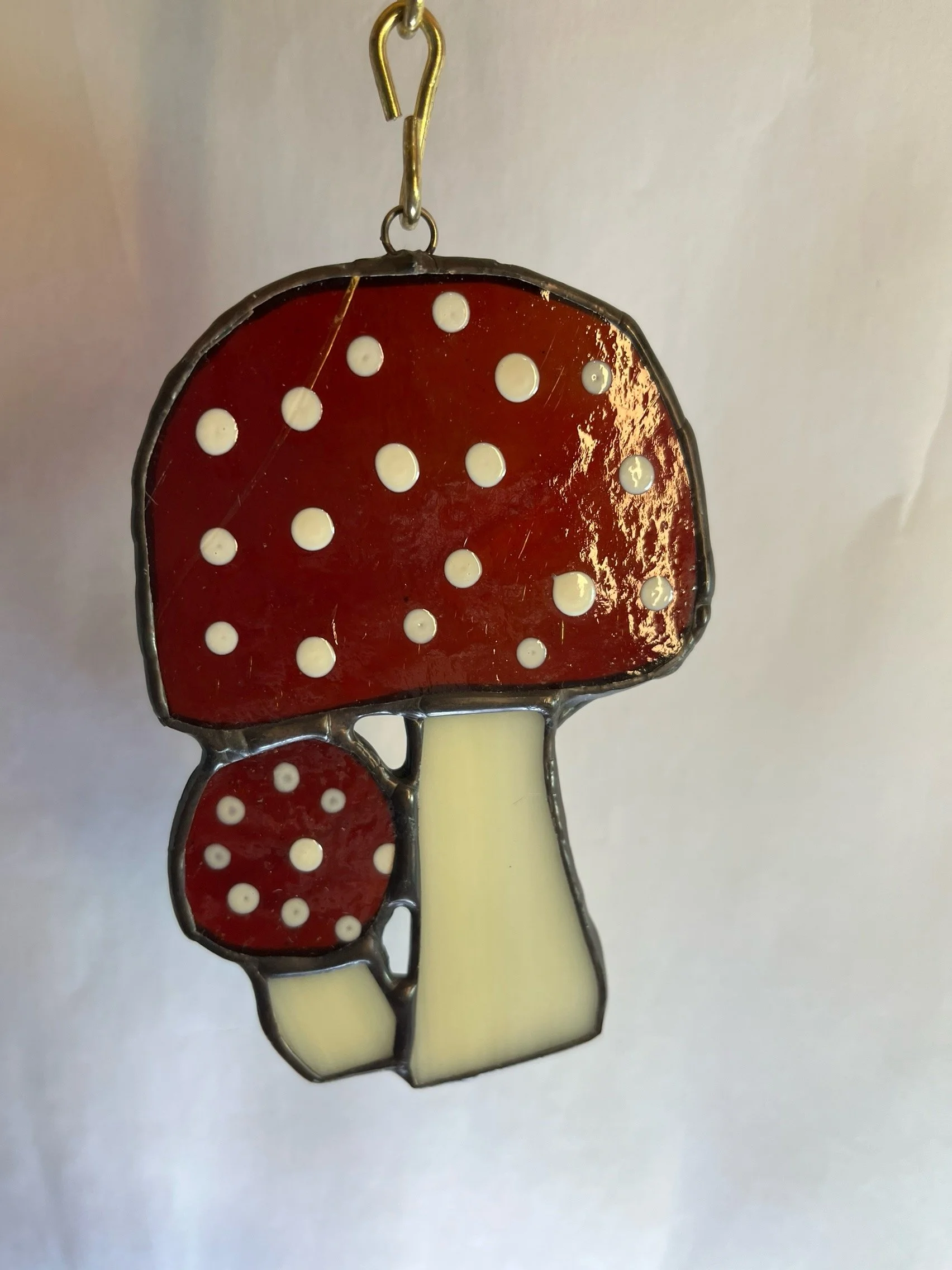 A stained glass sun catcher of two red mushrooms with white spots and cream-colored stems, hanging from a hook.
