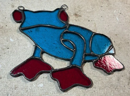 Colorful stained glass piece in the shape of a tree frog, with blue body and red eyes and feet, on a light surface.