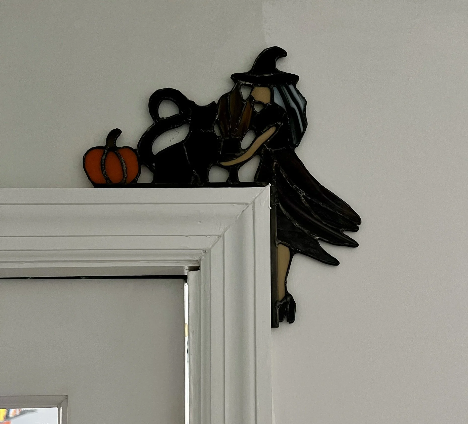 Stained glass Kitchen Witch and a pumpkin on a white wall near a white door frame.
