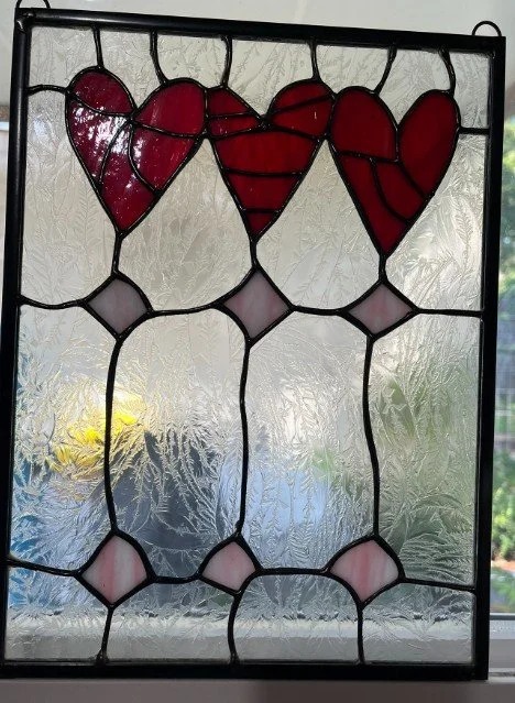 Stained glass window with three red hearts at the top and decorative pink and clear glass shapes below, set against a textured background.