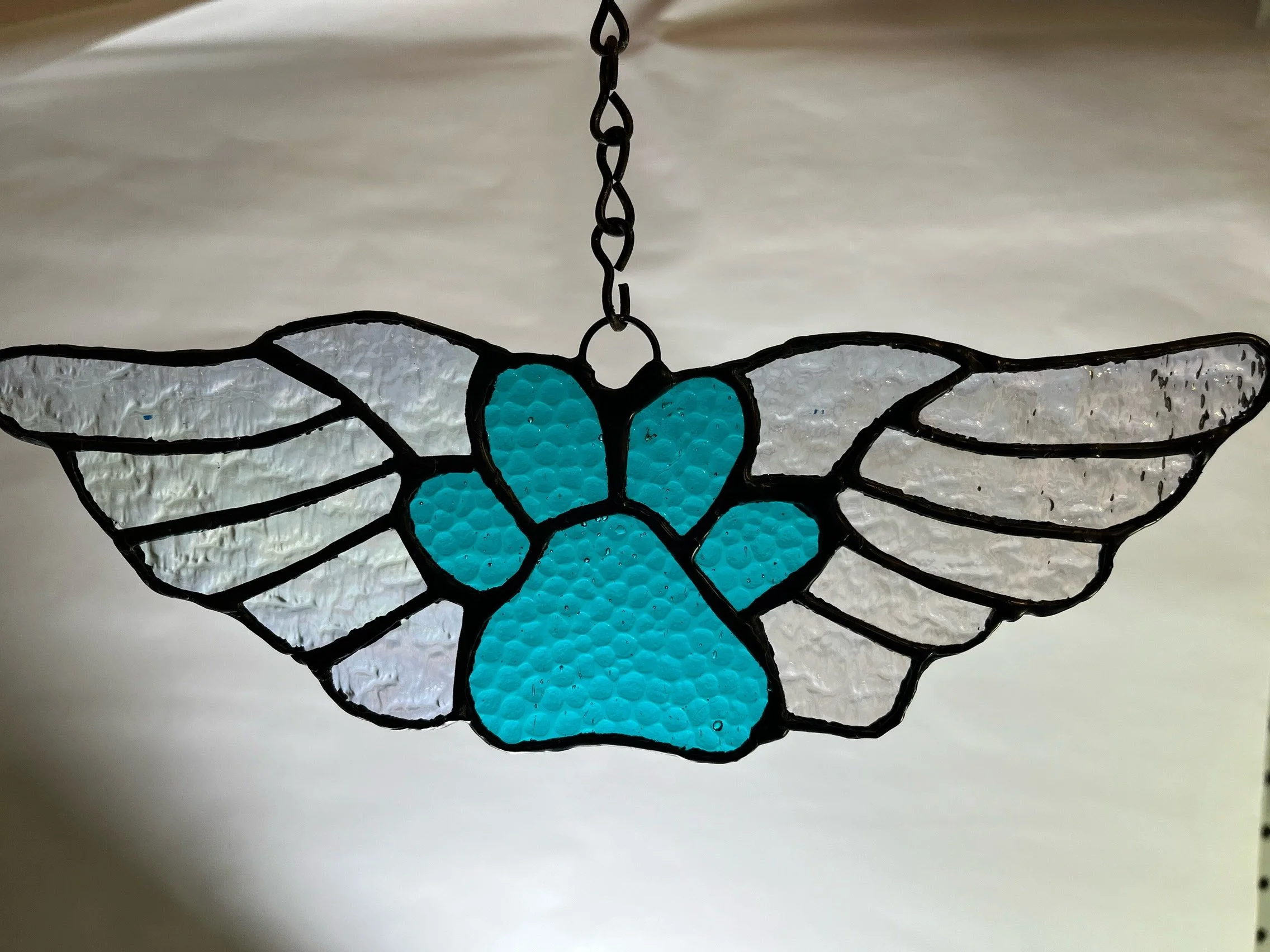 Stained glass sun catcher featuring a turquoise paw print with clear wings, hanging from a chain.