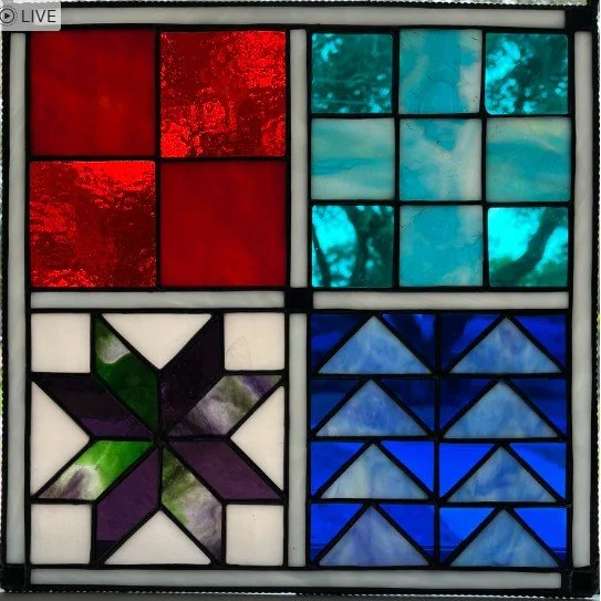 A colorful stained glass window divided into four quilt squares, each with different geometric patterns in red, turquoise, green, purple, and blue.