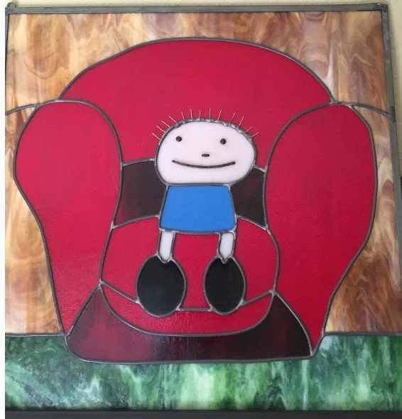 Colorful stained glass window of a cartoon character sitting in a red armchair. The character has a white face, black dot eyes, a small black mouth, and is wearing a blue shirt and black shoes. The background includes wooden textures and green at the