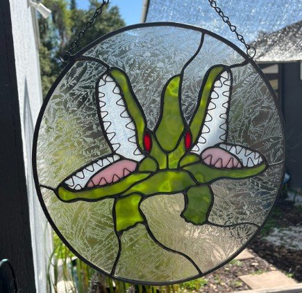Seminole Heights Two Headed Gator stained glass window with green and red accents, hanging outdoors with sunlight shining through.
