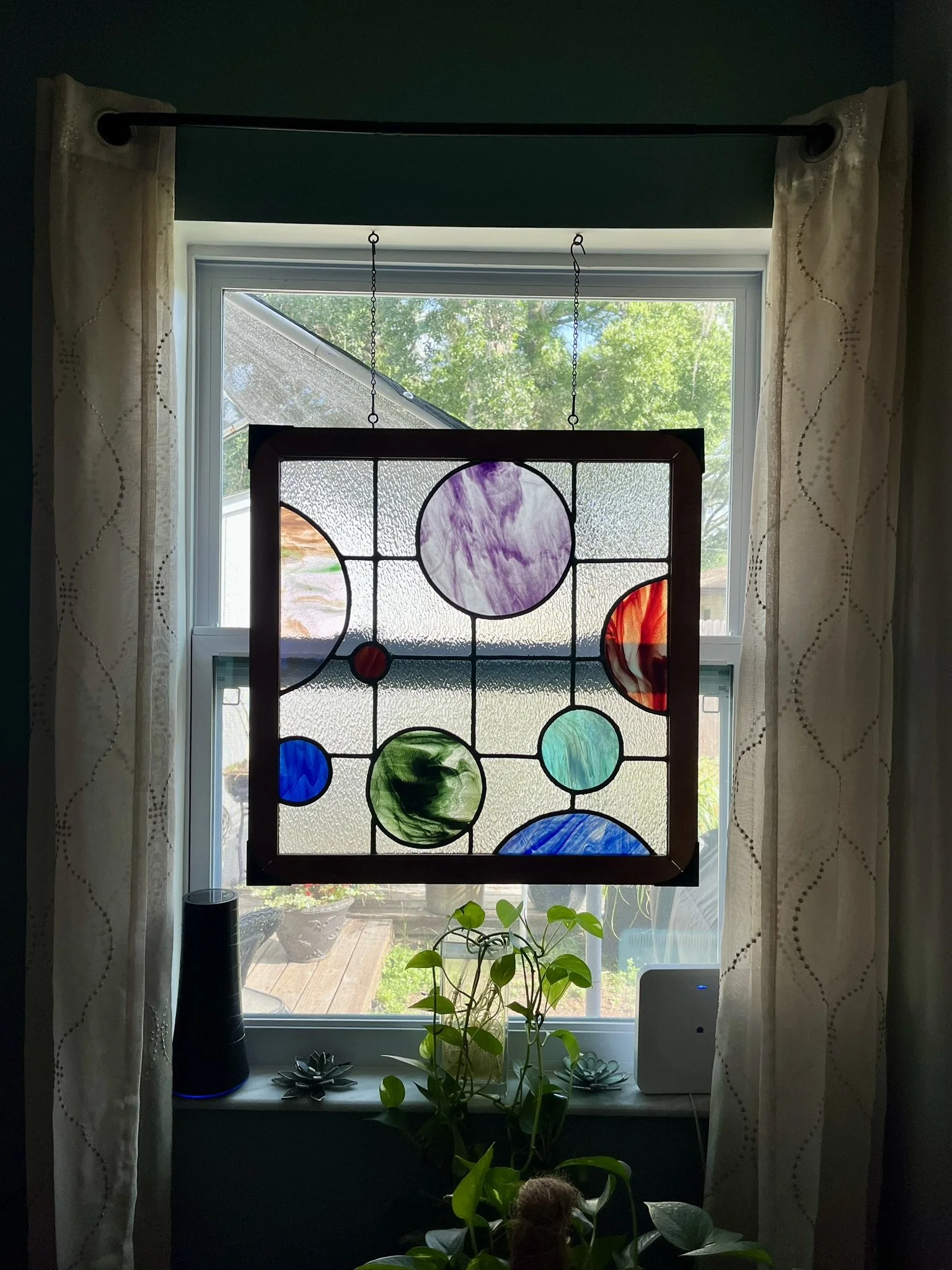 Decorative stained glass window hanging in front of a window, with a garden and trees outside.