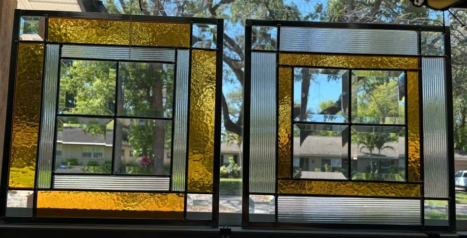 Two geometric stained glass window panels with colored and textured glass, placed against a window with trees and houses visible outside.