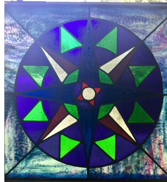 A colorful stained glass Mariner's Compass featuring blue, green, purple, white, red, and black sections.