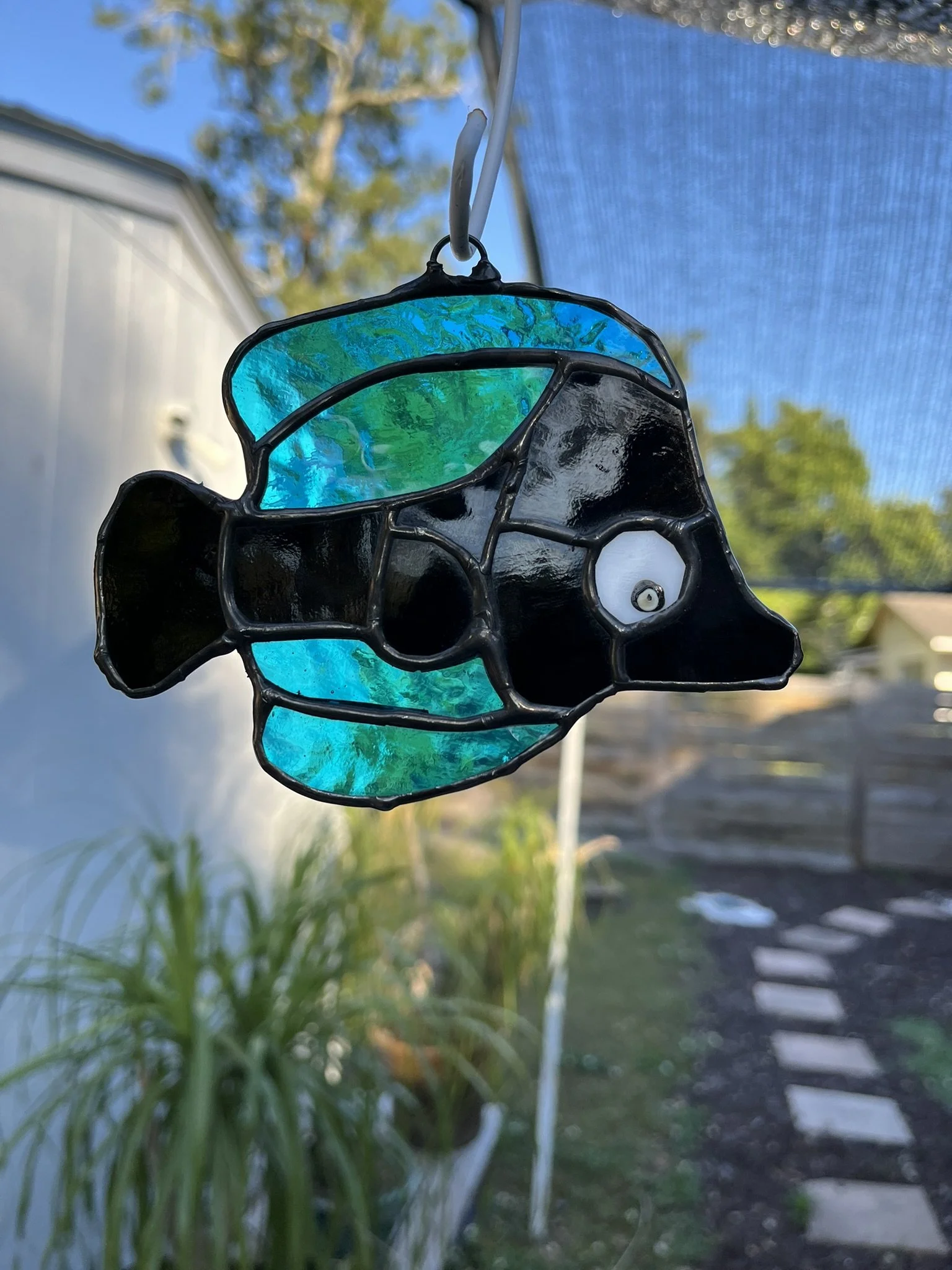 Colorful stained glass fish sun catcher hanging outdoors with a garden and pathway in the background.