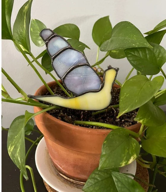 A potted plant with green leaves and a snail-shaped stained glass decoration in the soil.